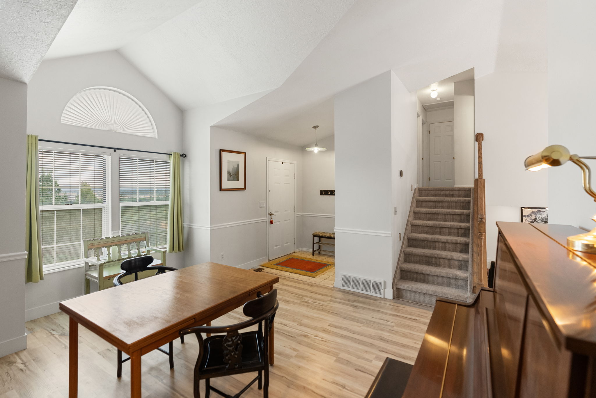 Vaulted ceiling with an arched window above a double window, wood-finish flooring, a carpeted staircase with wood railing, and a white entry door