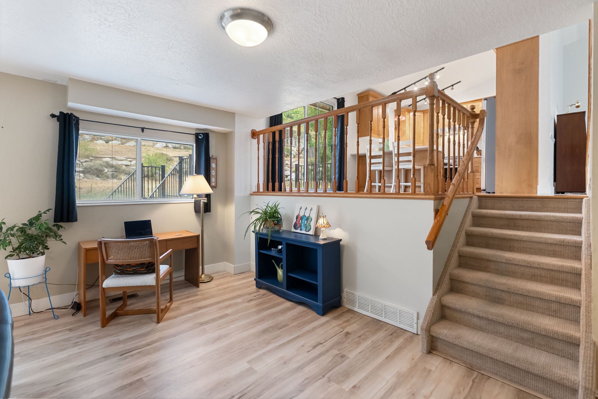 Spacious lower-level area with wood-finish flooring, a large window, a carpeted staircase, and a wood railing overlooking an upper space