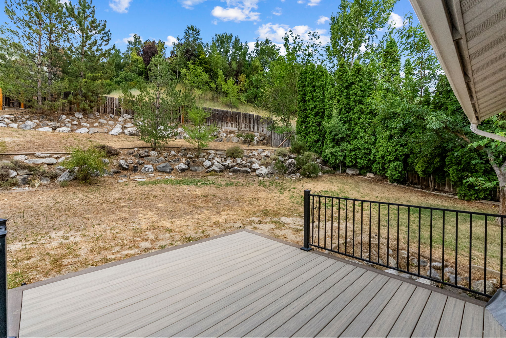 Outdoor deck space featuring composite decking, black metal railing, and a sloped backyard with mature trees and rock landscaping