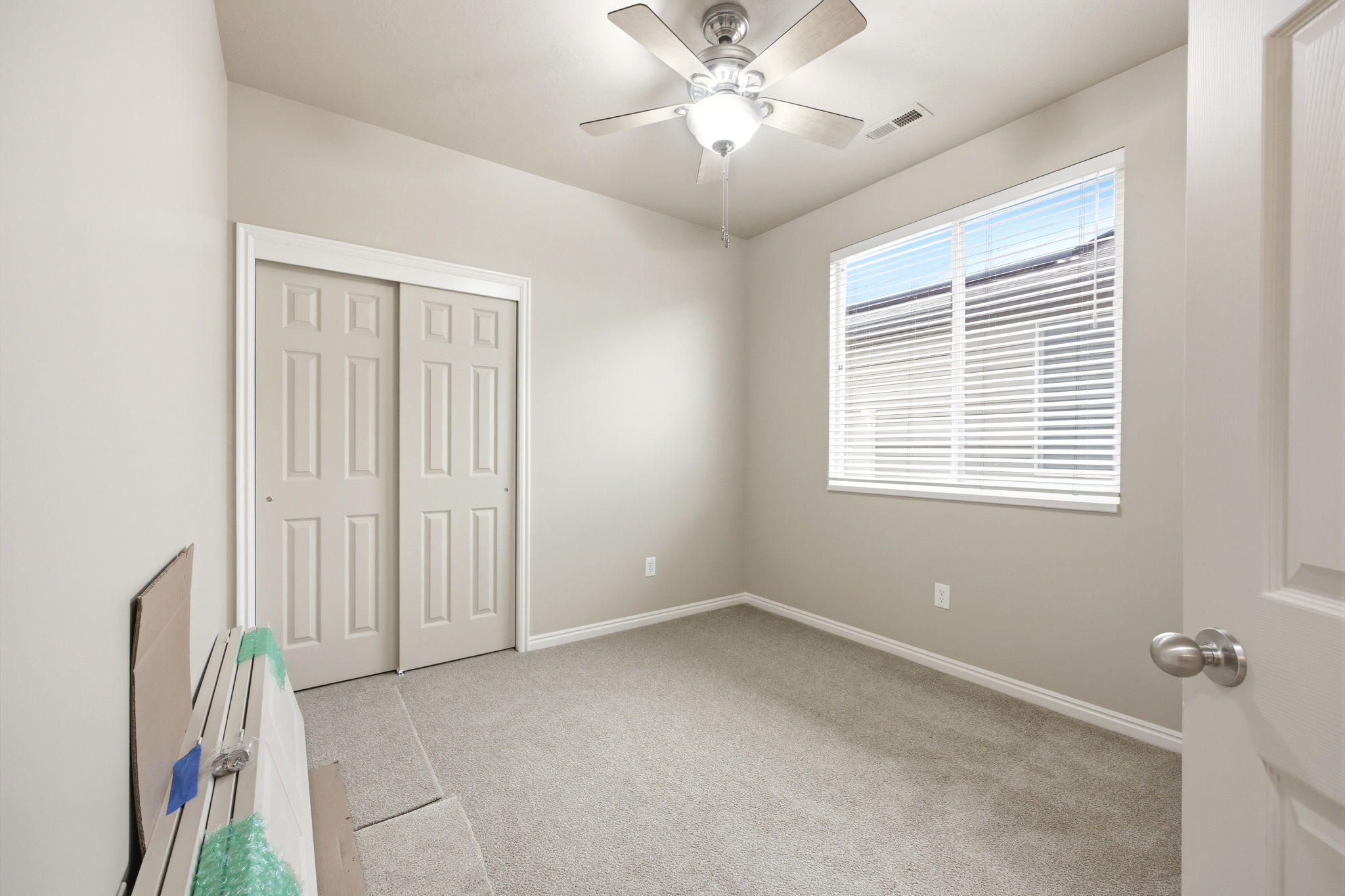 Neutral-toned room featuring a ceiling fan with light fixture, large window with blinds, and light gray wall-to-wall carpeting