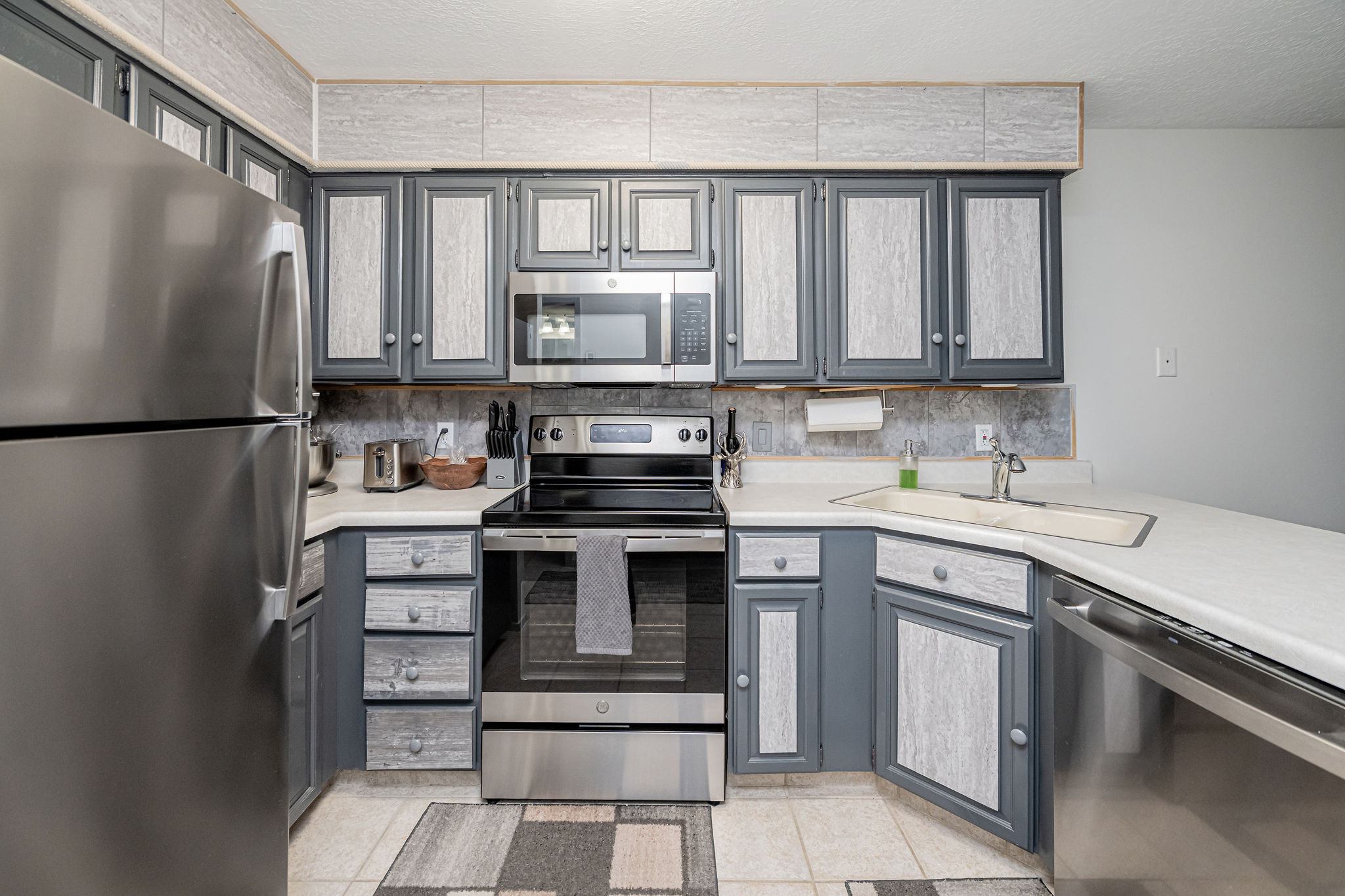 Kitchen featuring stainless steel appliances, grey and light wood-finish cabinetry, a corner sink with a stainless steel faucet, white countertops, and light tile flooring