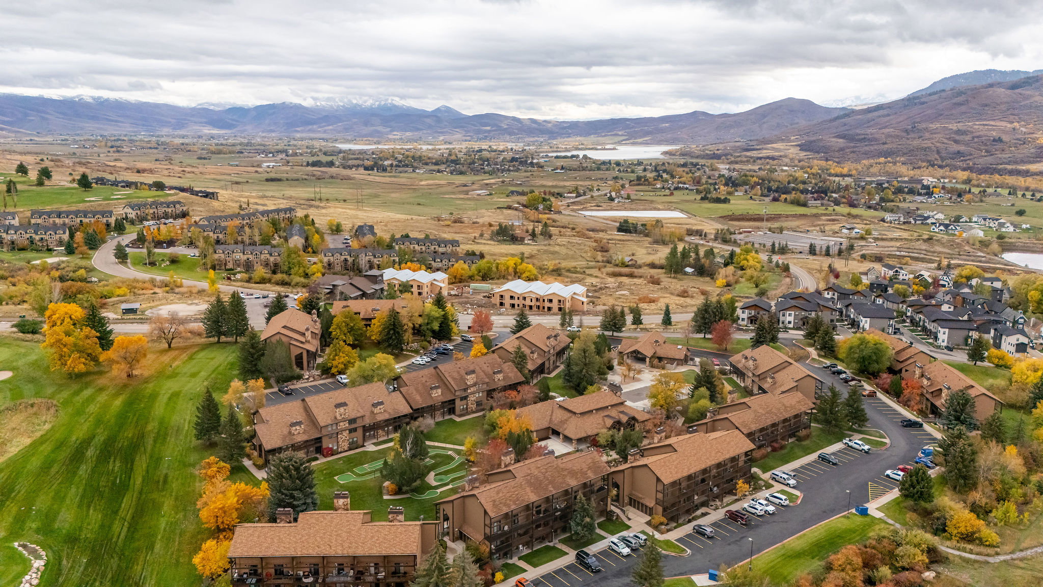 Expansive aerial capture of a residential development featuring multiple buildings with brown roofs, surrounded by mature trees displaying autumn foliage