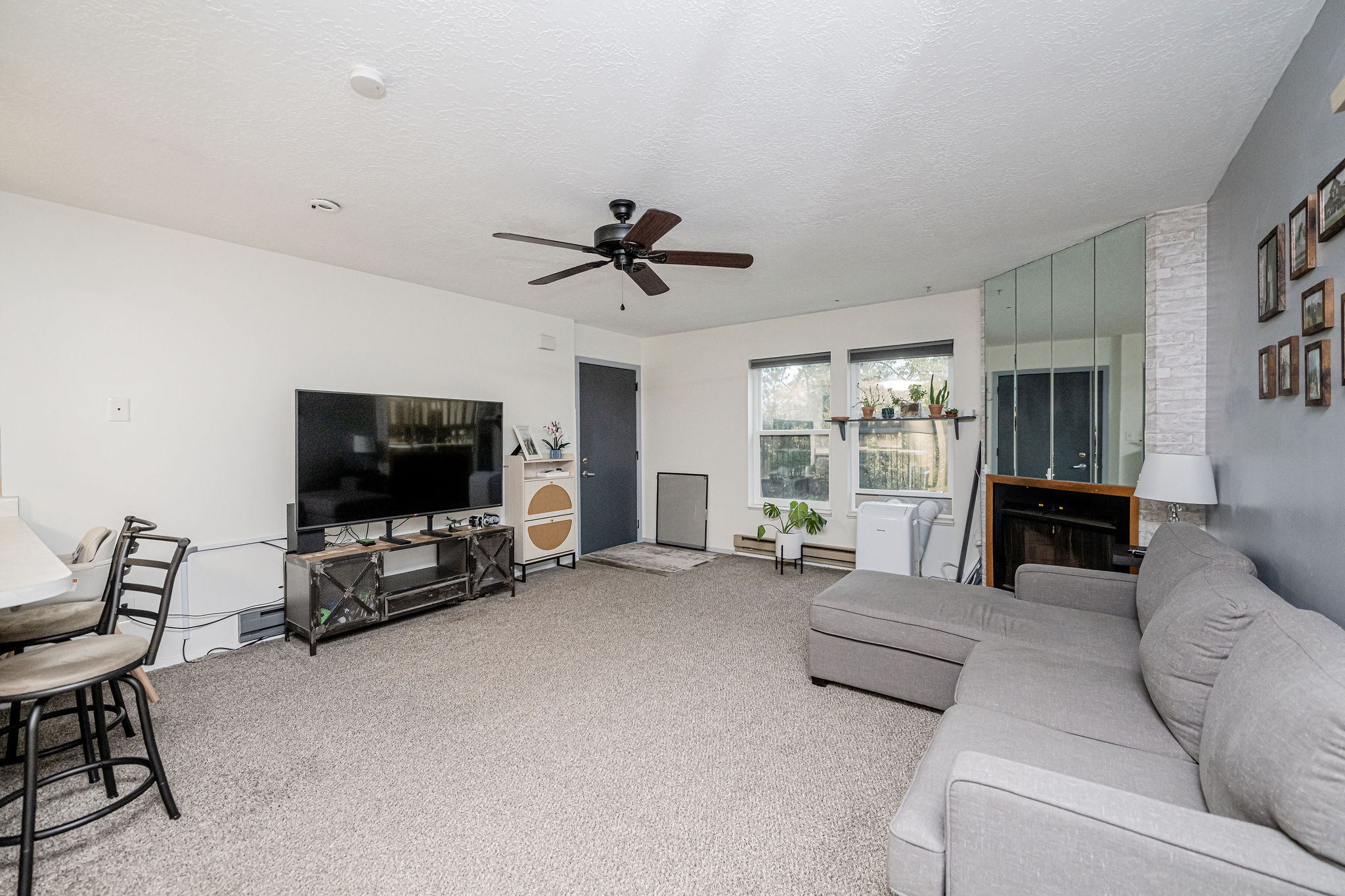 Spacious living area featuring light gray carpeting, a dark ceiling fan, multiple windows with natural light, a mirrored wall accent, and a built-in electric fireplace with a wooden mantel