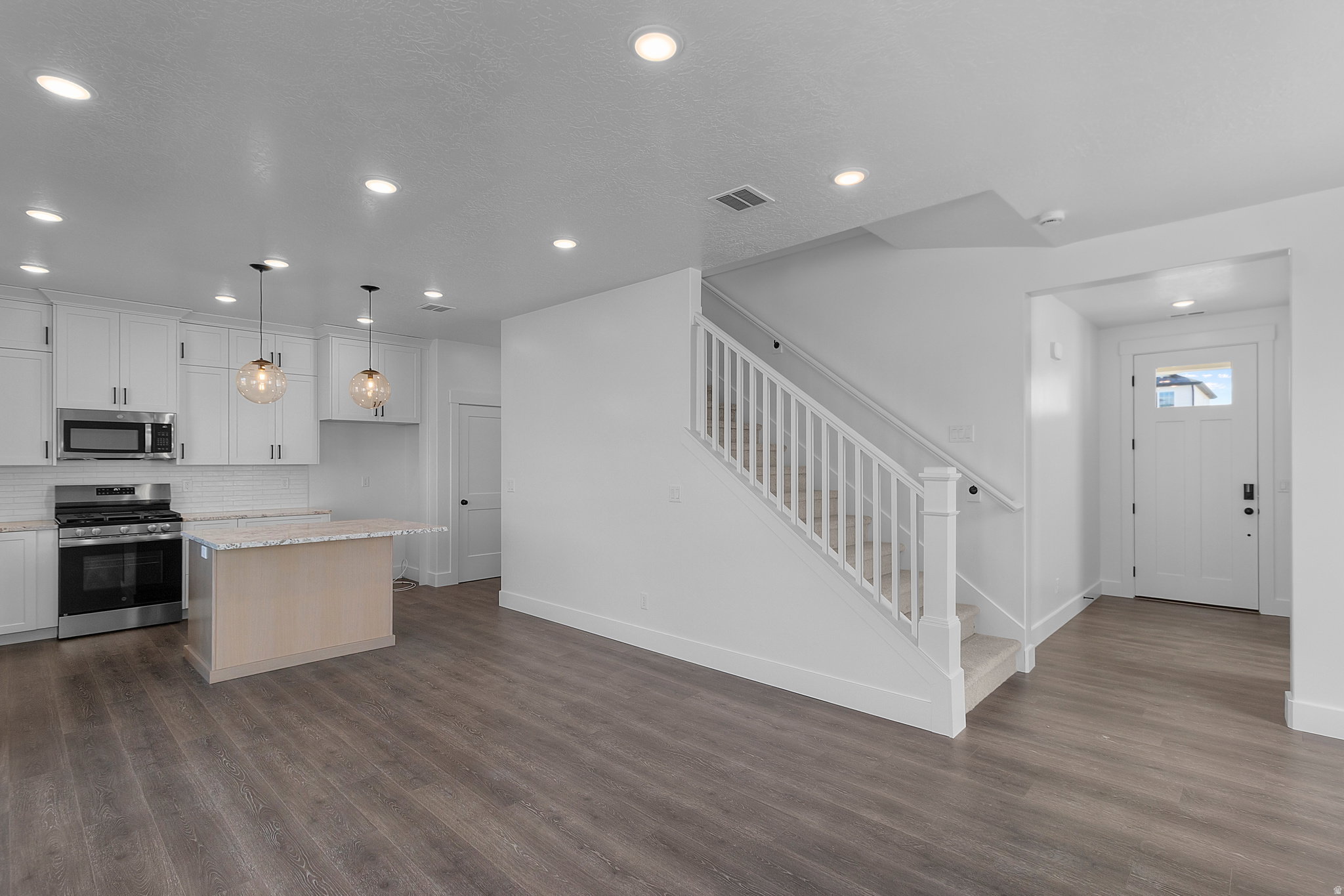 Open-concept living area featuring wood-finish flooring, a contemporary kitchen with white cabinetry and stainless steel appliances, a central island, a carpeted staircase with white railings, and recessed lighting throughout
