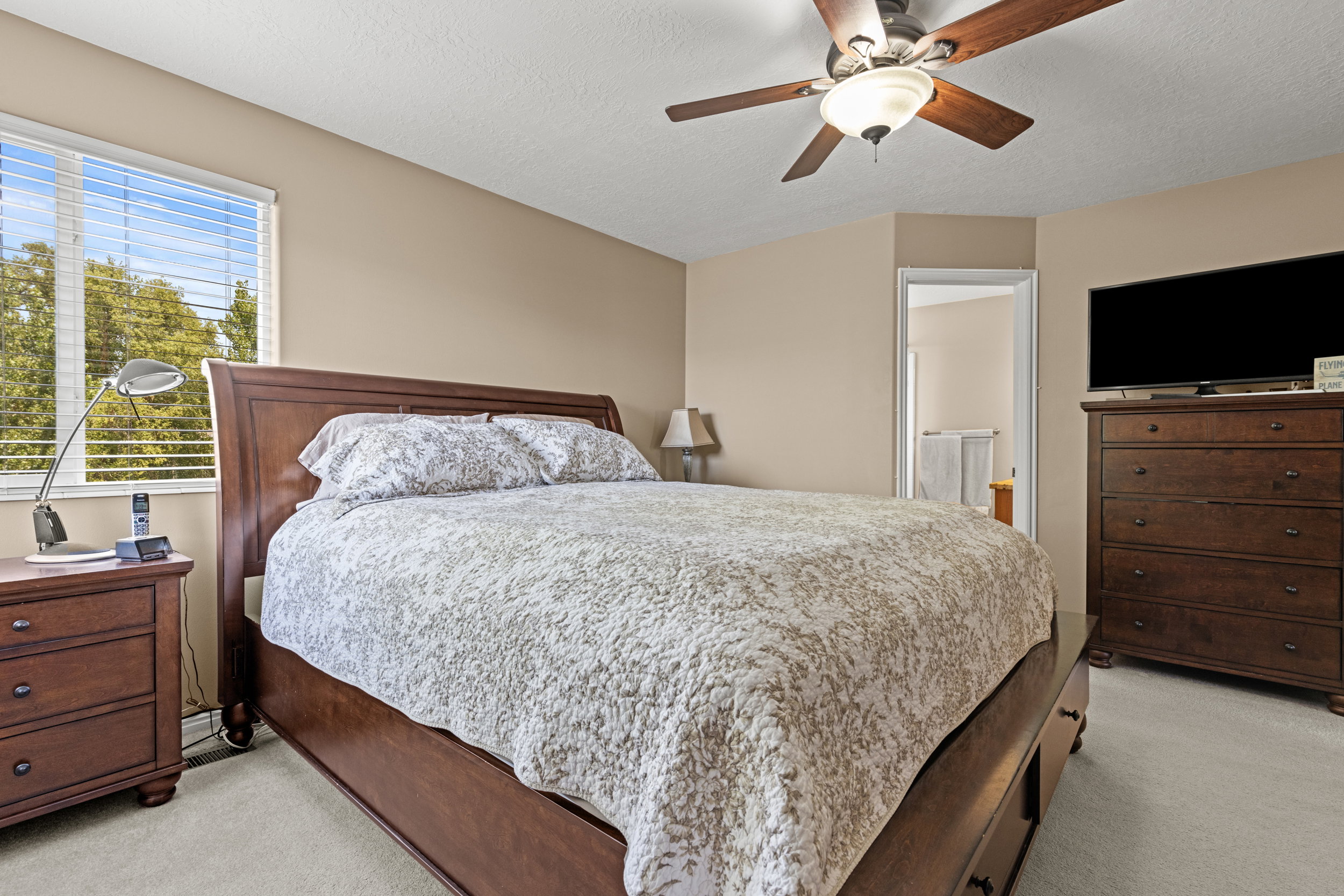 Carpeted bedroom featuring a large window with horizontal blinds, a ceiling fan with integrated lighting, and direct access to a separate space