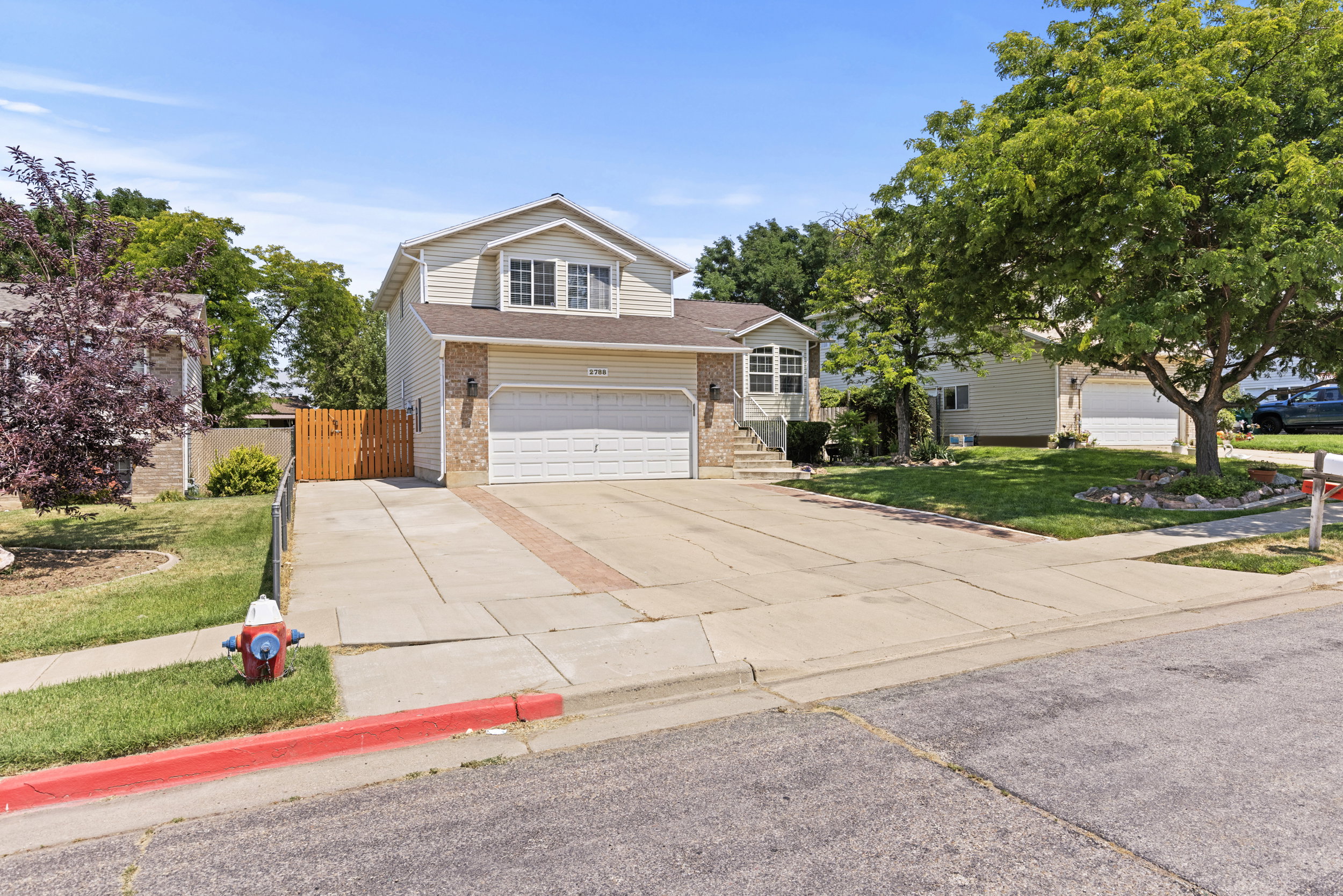 Two-story exterior with light-colored siding and brick accents, featuring a two-car garage, mature landscaping, a wooden privacy fence, and a concrete driveway with a brick border