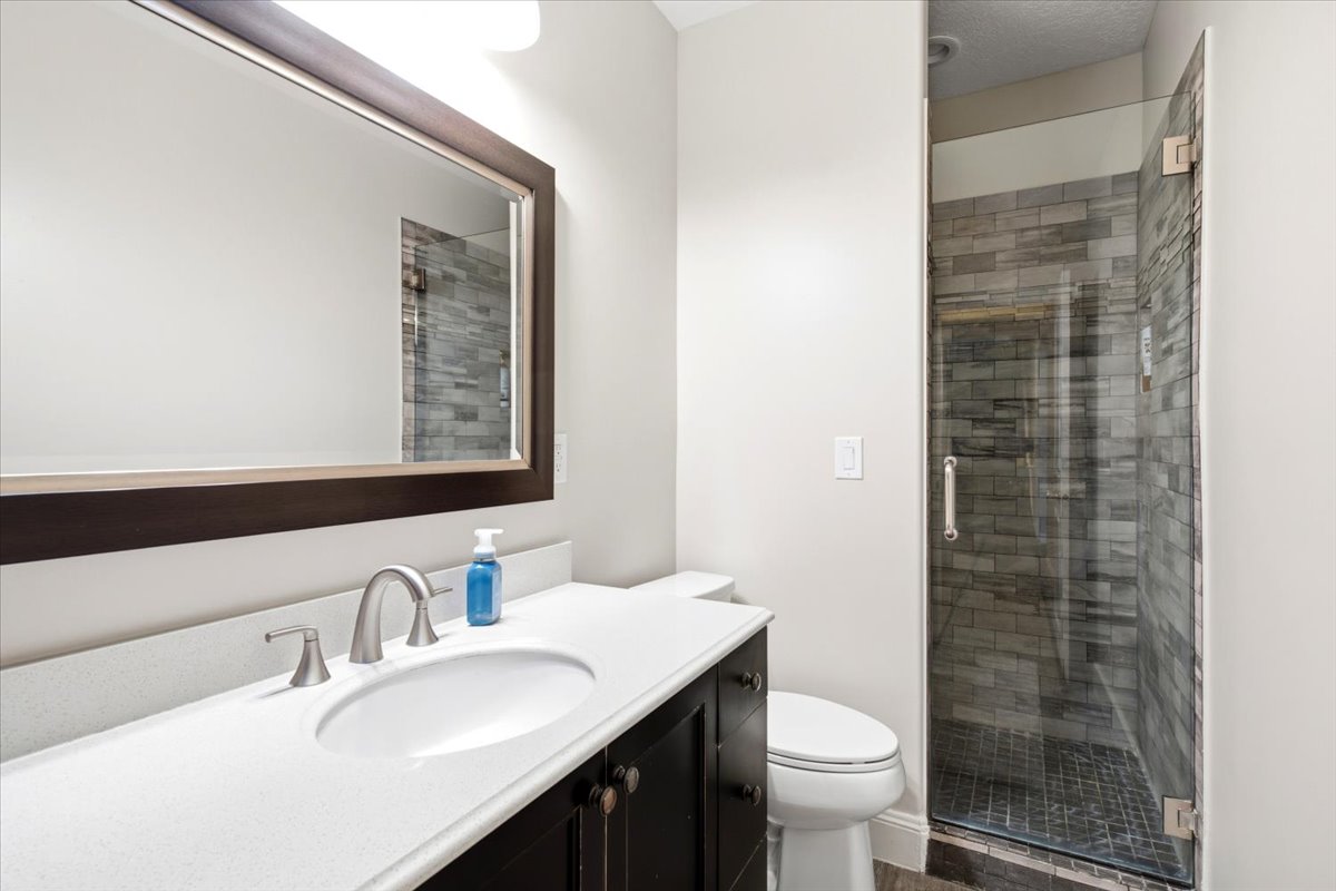 Bathroom featuring a dark wood-finish vanity with a white countertop and undermount sink, a framed mirror, and a glass-enclosed shower with stacked stone-look wall tile