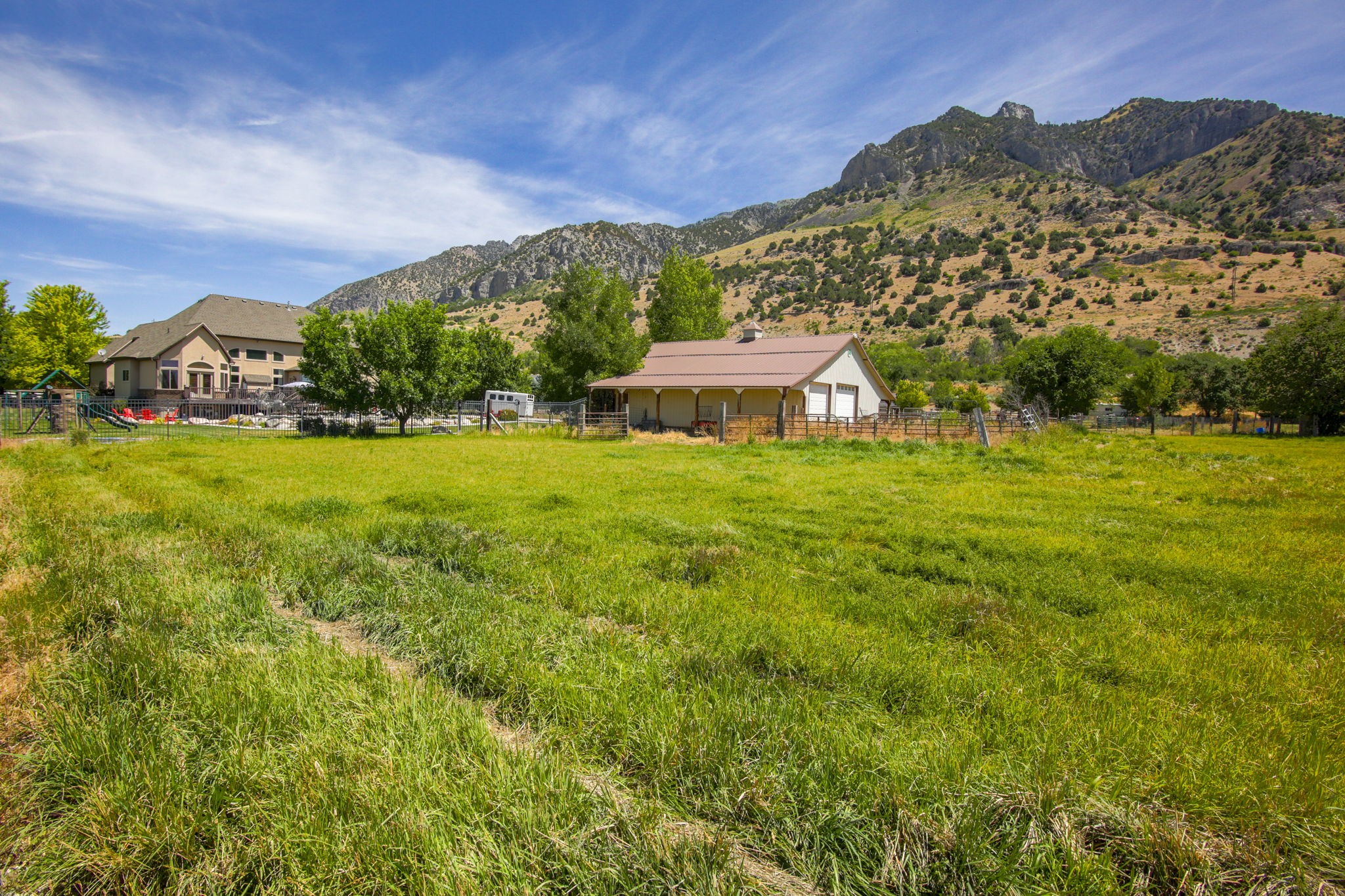 Expansive green field bordering a detached structure with a brown roof and two garage doors, set against a backdrop of a mountainous landscape