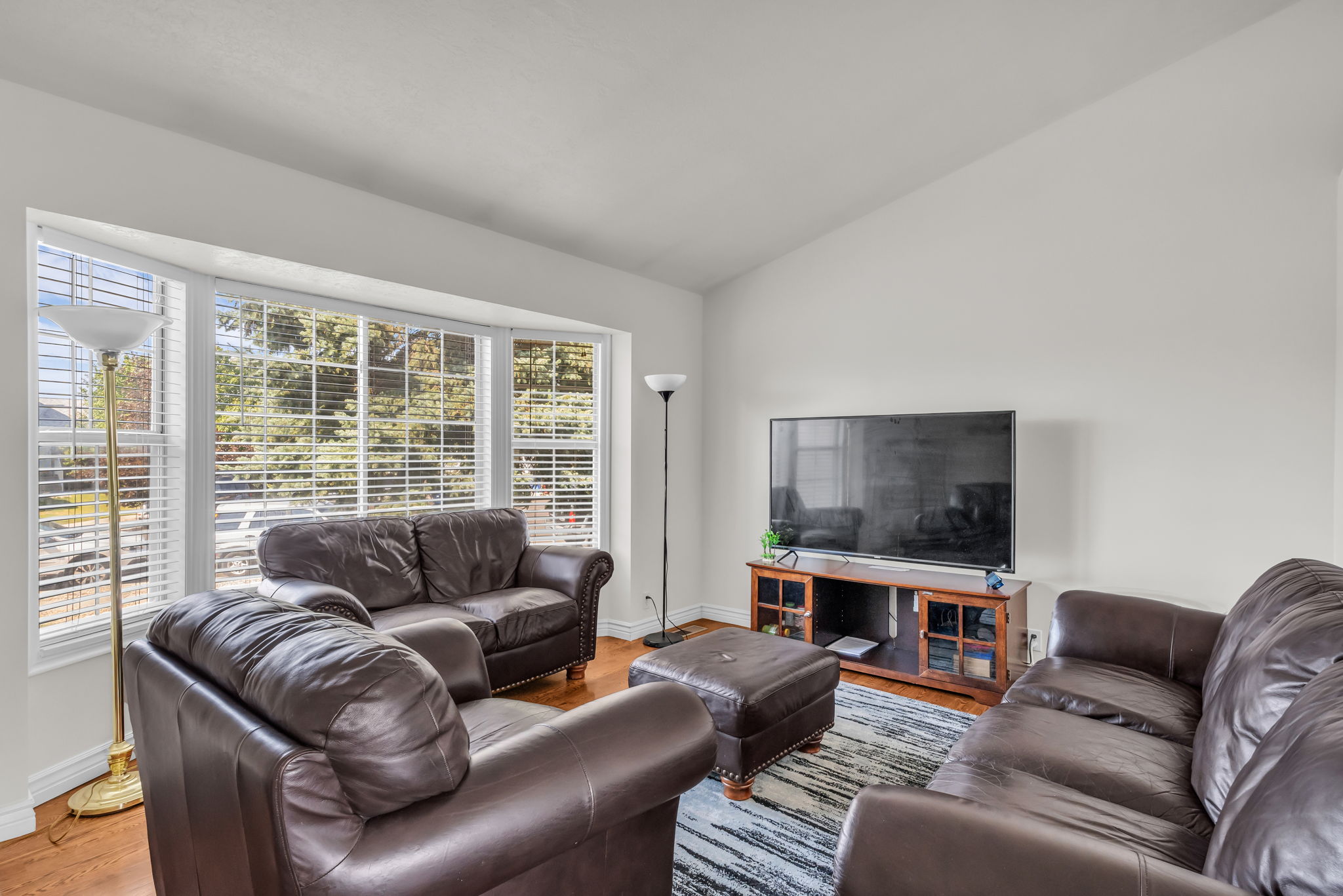 Living area featuring a prominent bay window with white blinds, vaulted ceiling, and wood-finish flooring