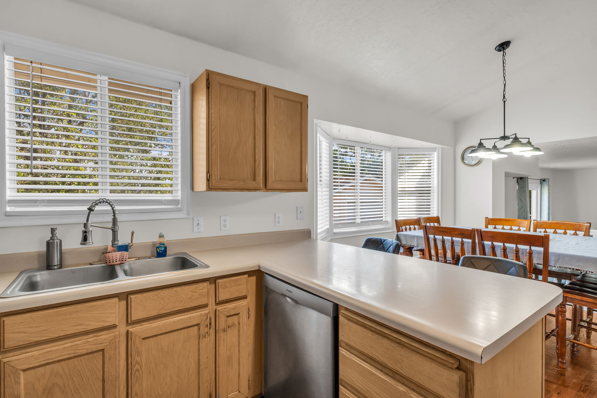 Kitchen featuring a double stainless steel sink with a high-arc faucet, wood-finish cabinetry, light-toned countertops, and a built-in dishwasher