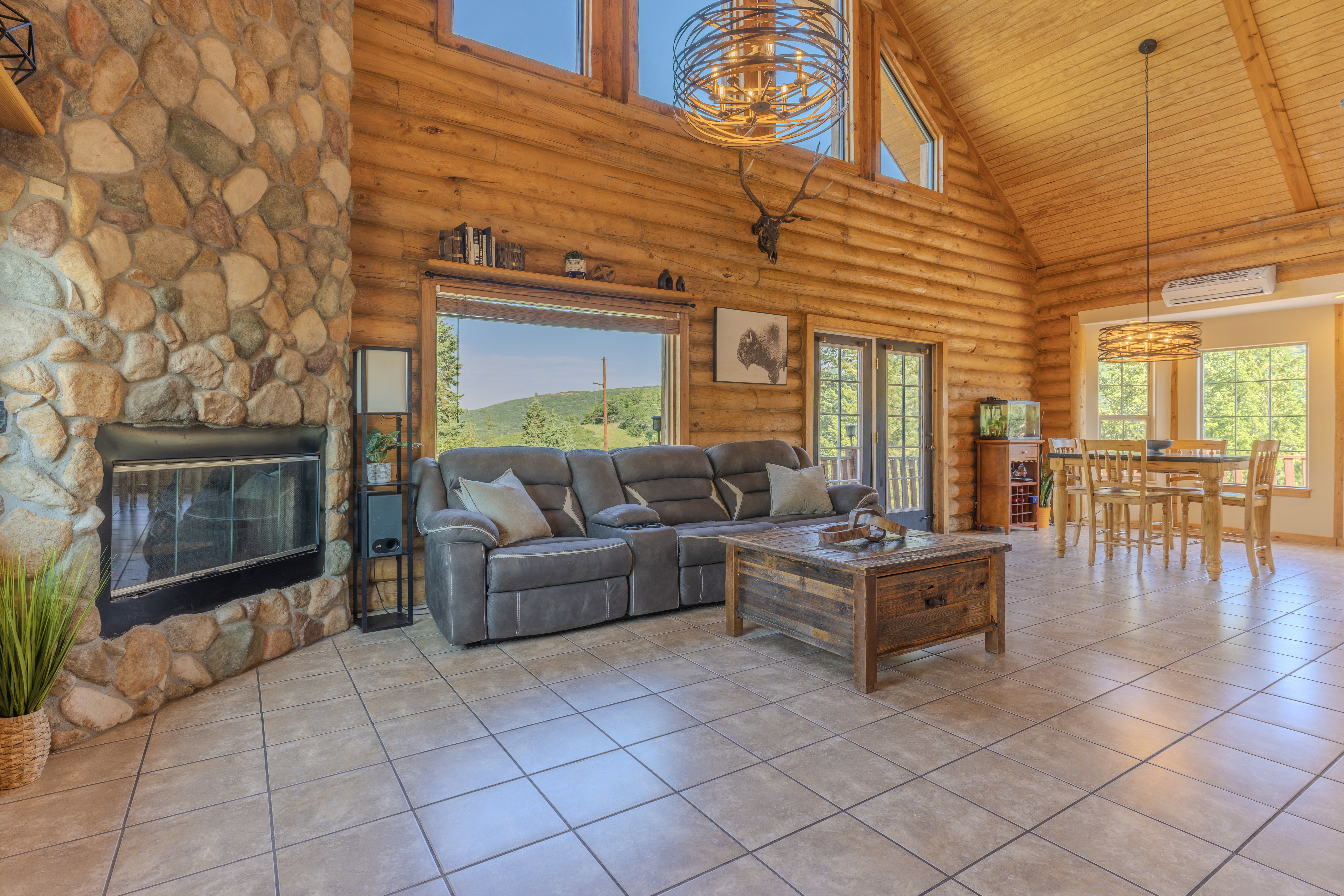 Open-concept living space featuring a floor-to-ceiling river rock fireplace, wood-finish walls and vaulted ceilings, large picture windows, and tile flooring