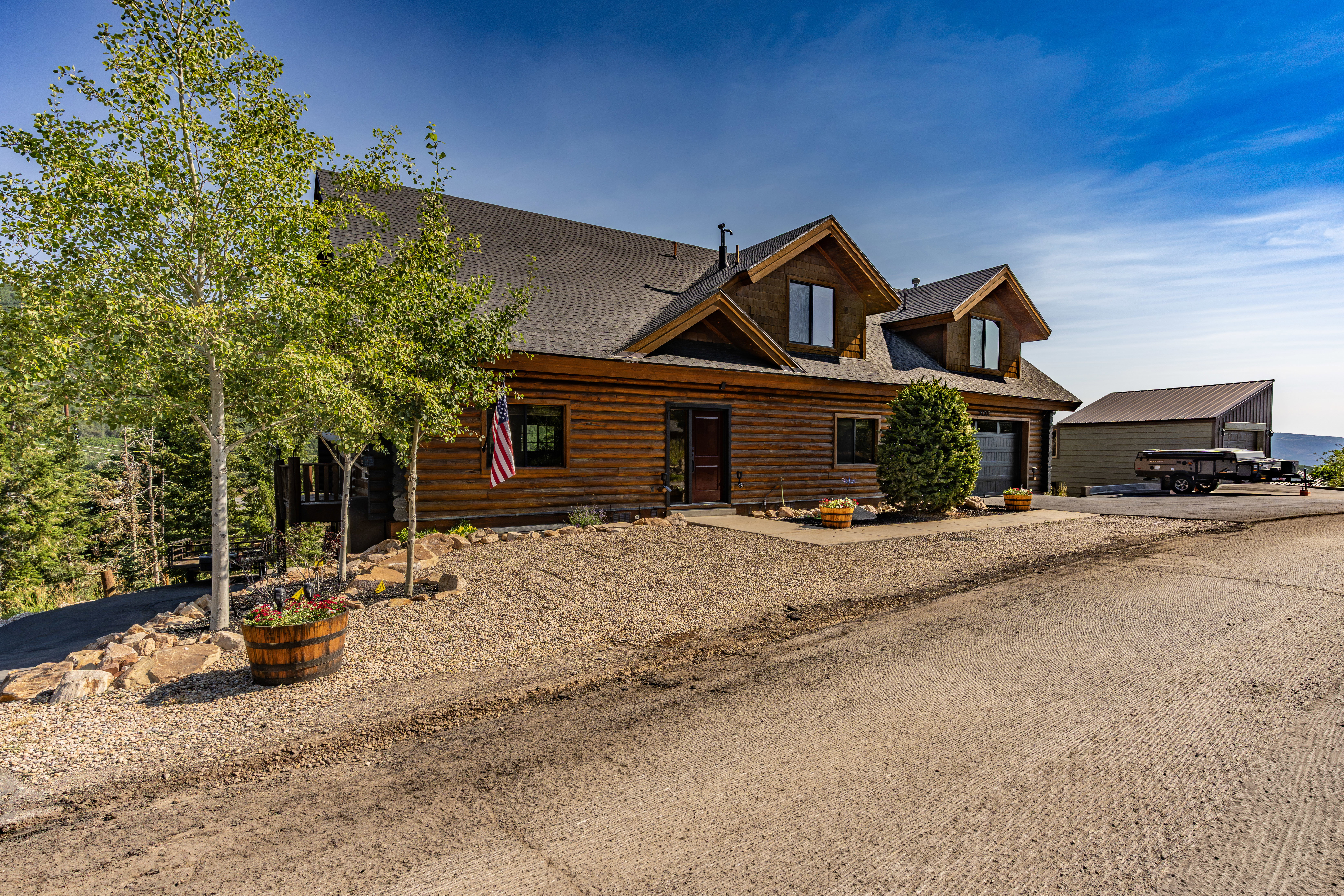 Log cabin exterior featuring a dark asphalt shingle roof, dormer windows, and an attached two-car garage