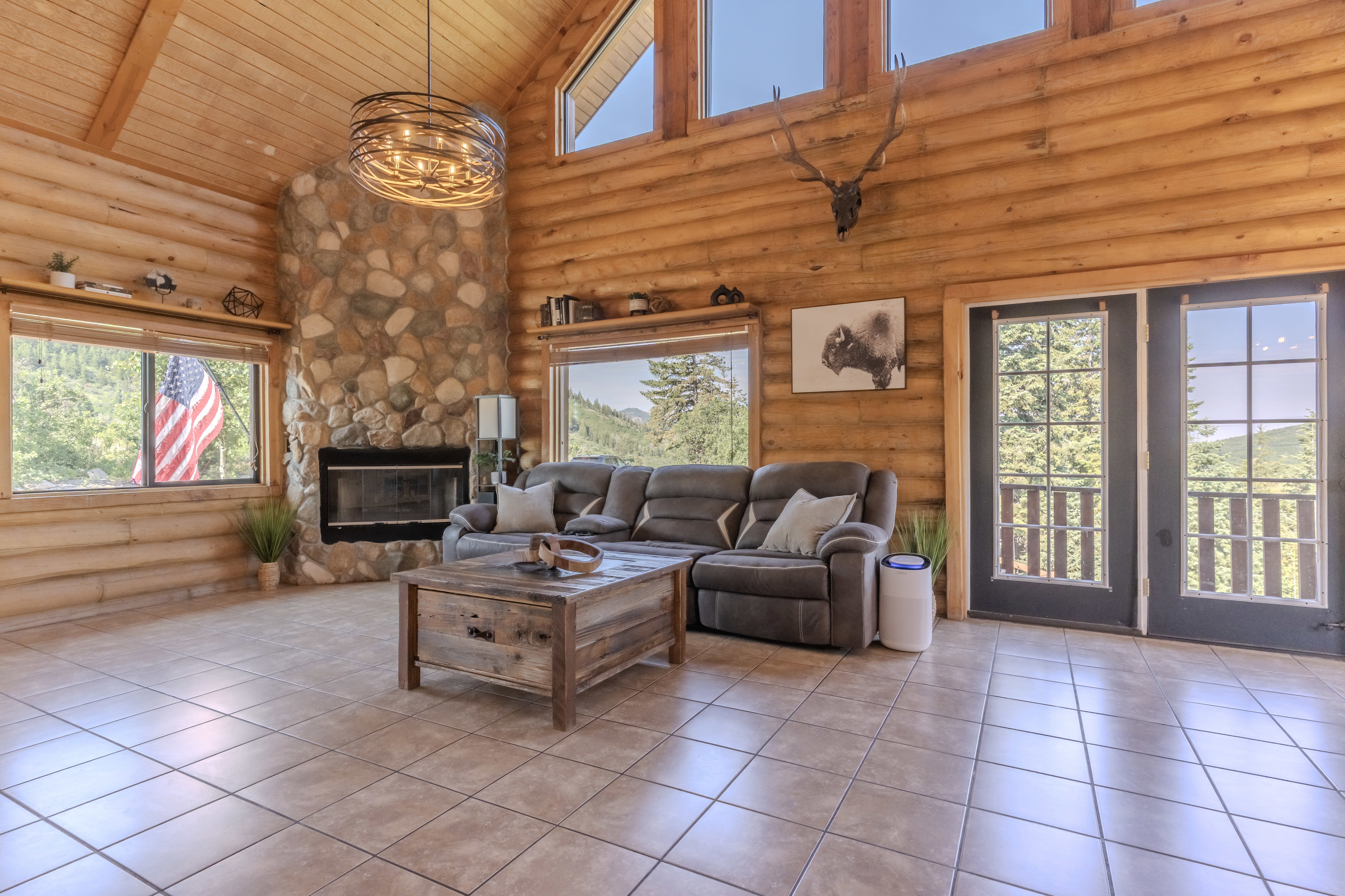 Vaulted ceiling great room featuring log walls, tile flooring, a stone fireplace, and double French doors opening to a balcony