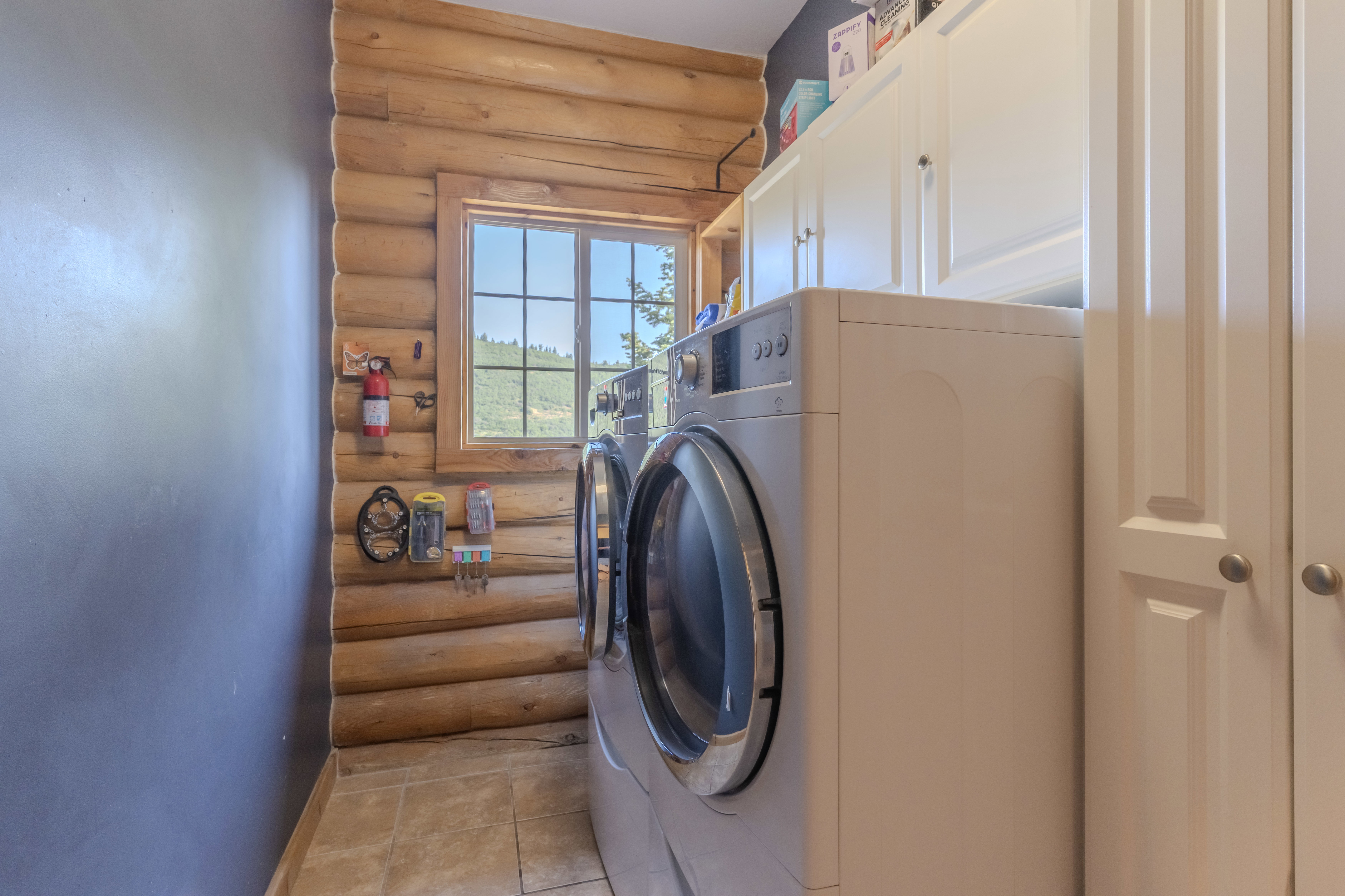 Laundry area featuring natural log walls, a window with exterior scenery, tiled flooring, and white cabinetry