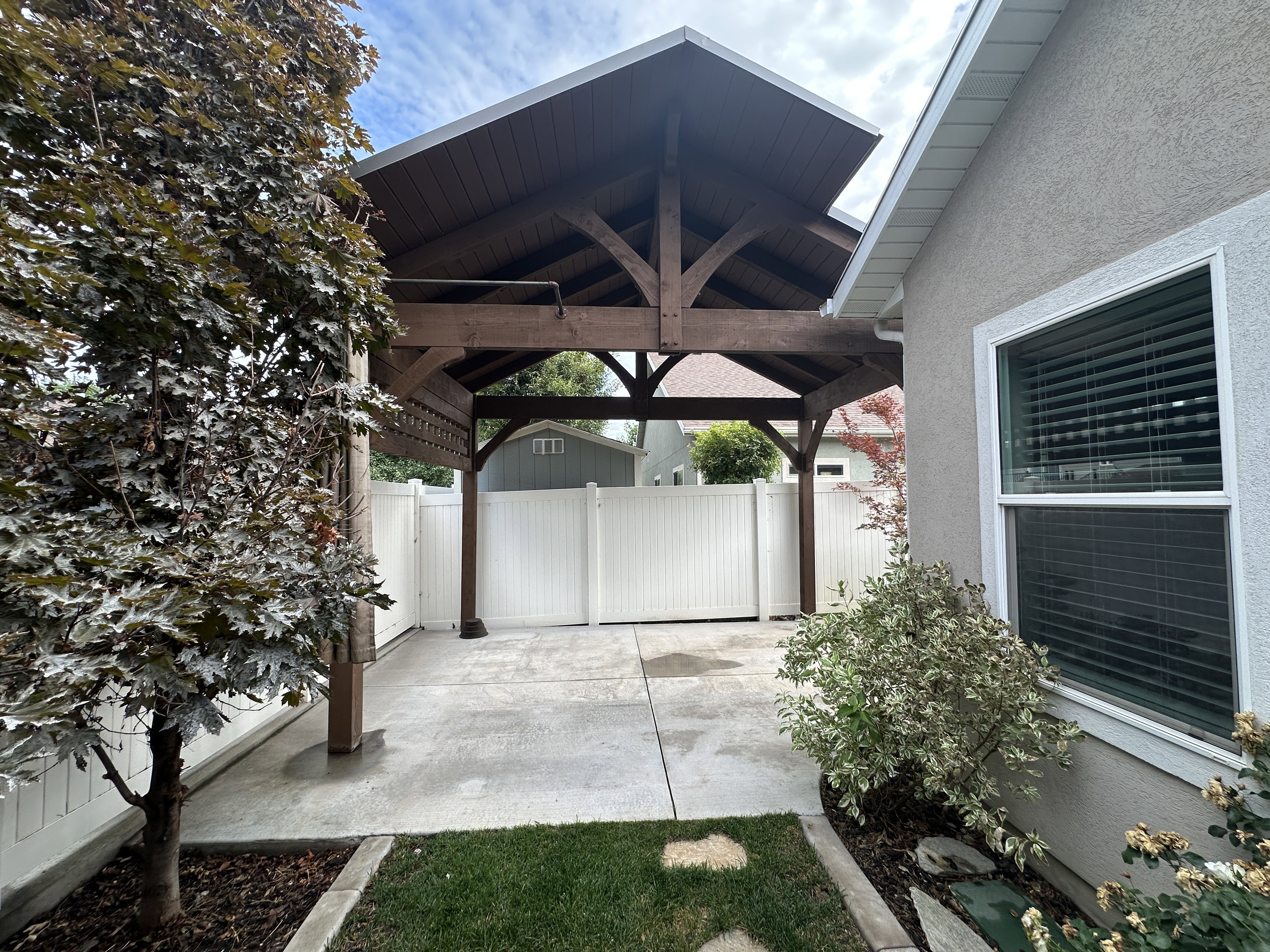 Wood-beam patio cover with a slatted roof, concrete patio, and white vinyl fencing