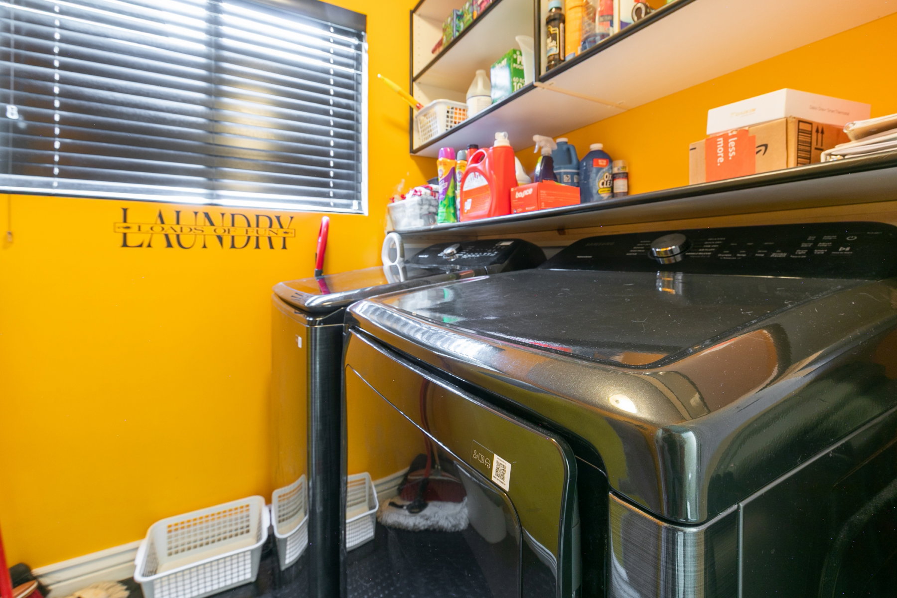 Dedicated laundry area featuring a window with horizontal blinds, overhead shelving, and a dark-toned washer and dryer set