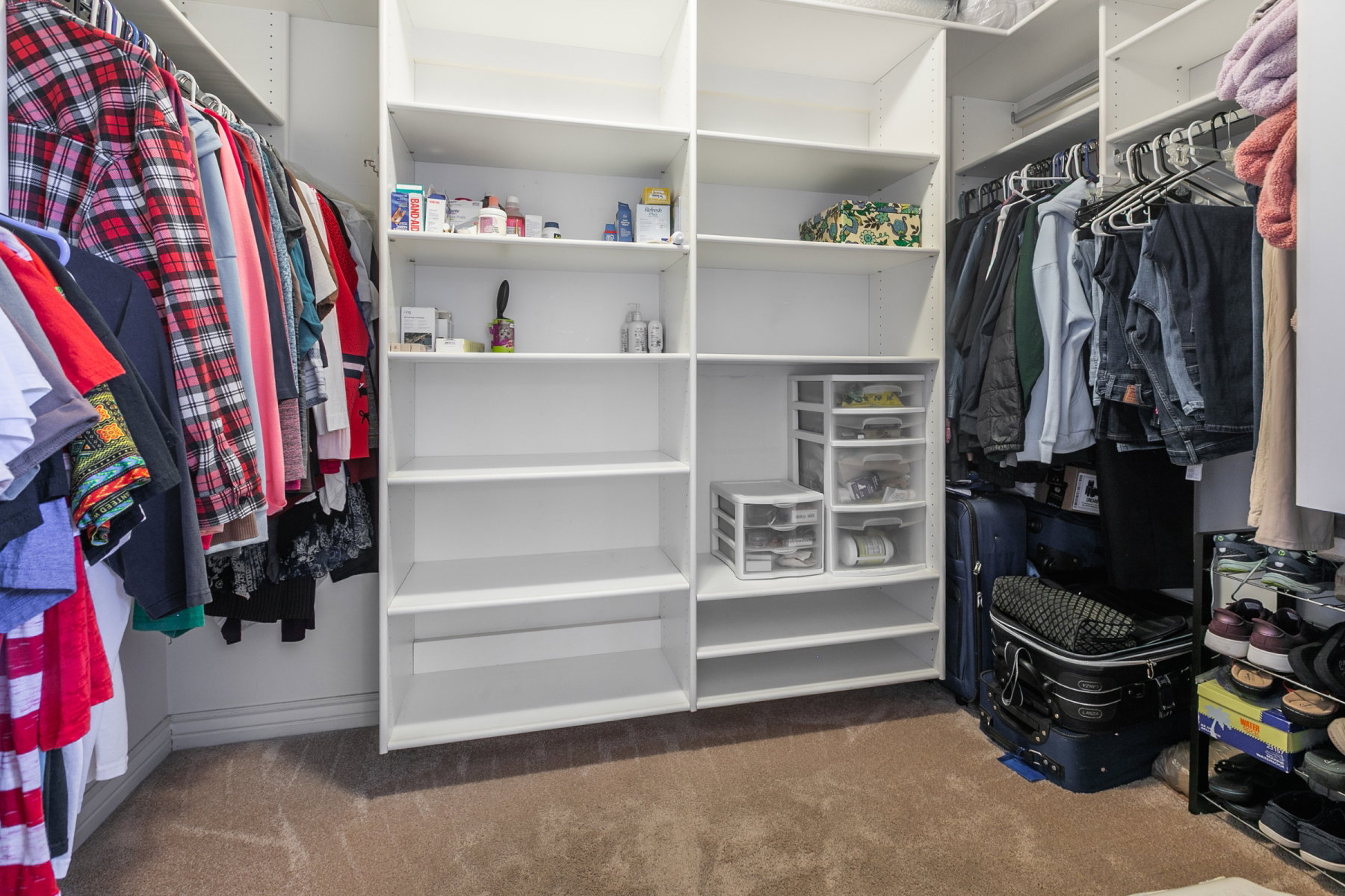 Spacious primary walk-in closet featuring built-in white shelving, multiple hanging rods, and carpet flooring