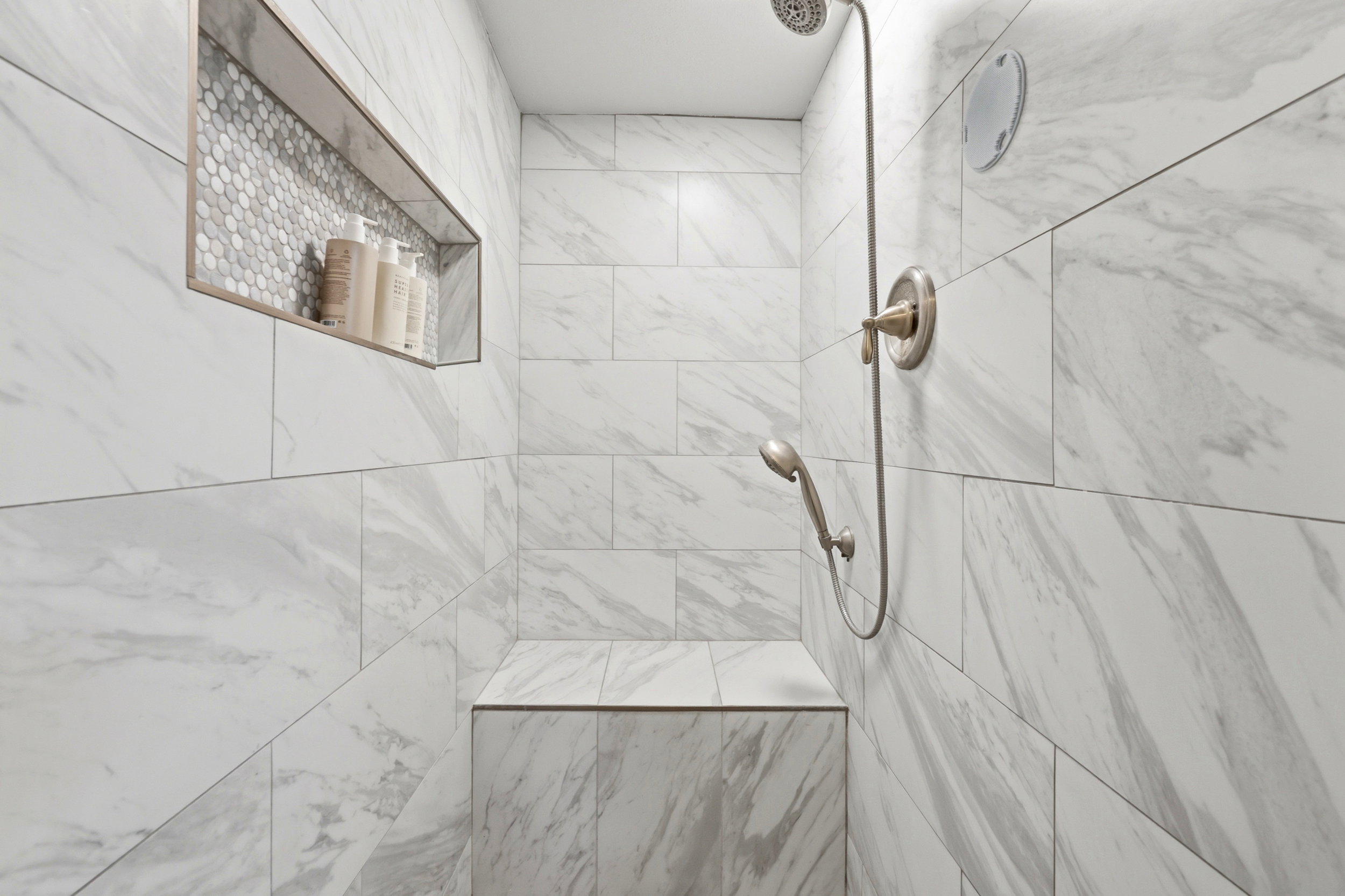 Tiled shower with a built-in bench, recessed niche featuring hexagonal mosaic tile, and a brushed nickel shower fixture with a handheld sprayer