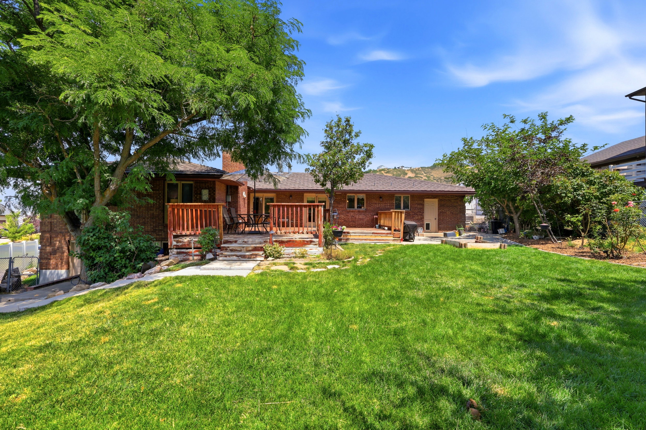 Backyard featuring a manicured lawn, multi-level wood deck, mature trees, brick exterior, and a visible chimney