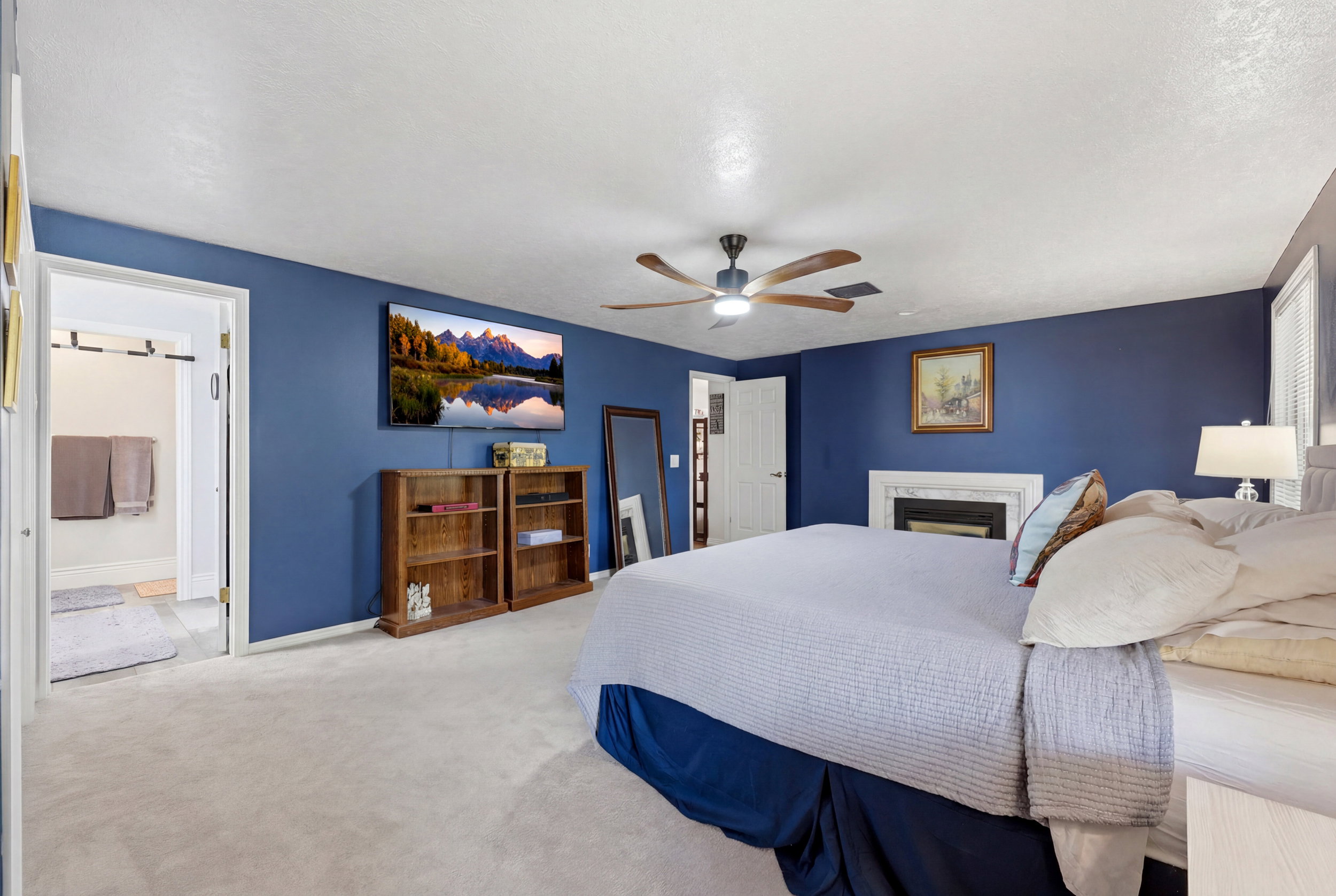 Carpeted room featuring a white fireplace with a dark insert, a ceiling fan with light, and an open doorway to an adjacent bathroom with light-colored flooring