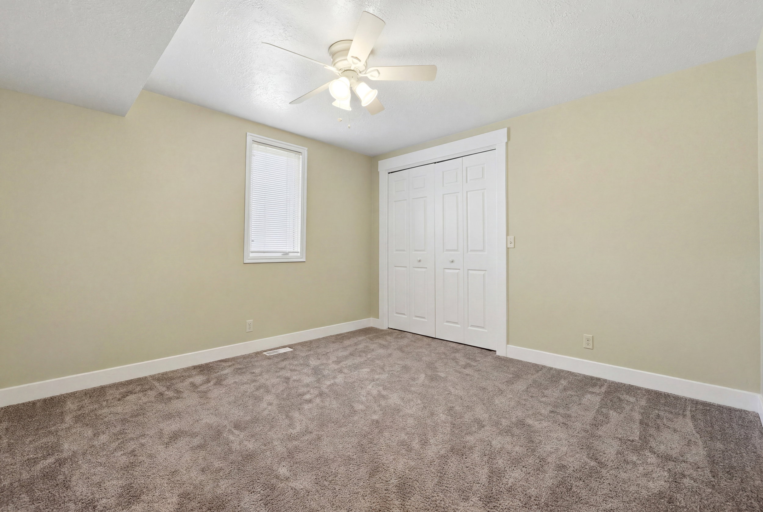 Carpeted room with neutral-toned walls, a ceiling fan with light fixture, a window with white trim, and a bi-fold closet with white doors