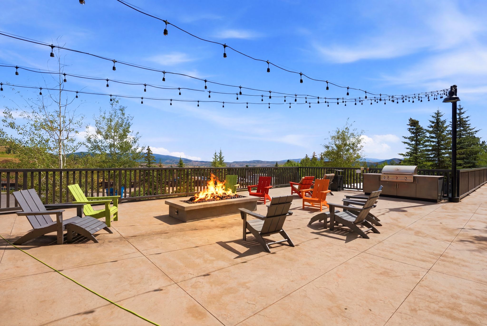 Expansive outdoor patio featuring a built-in fire pit, string lighting, integrated outdoor kitchen, and a surrounding wooden railing