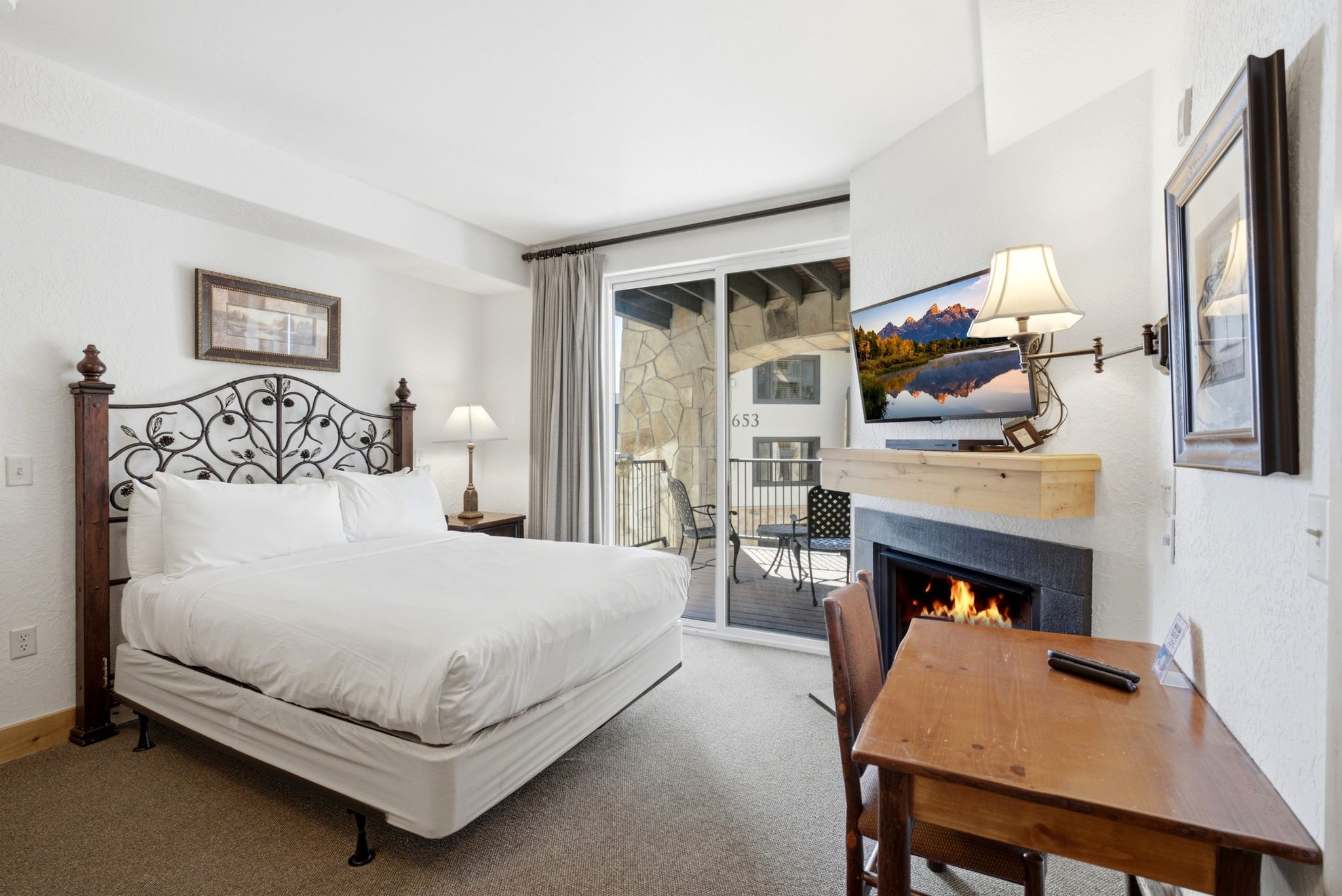 Comfortable sleeping area featuring a decorative wrought iron headboard, adjacent fireplace with a wood-finish mantel, and direct access to a private outdoor balcony with stone-finish exterior