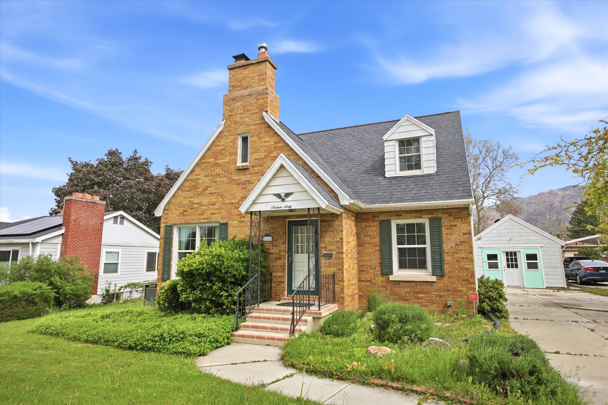 Brick facade home featuring a prominent chimney, dark shingle roof, dormer window, and green window shutters