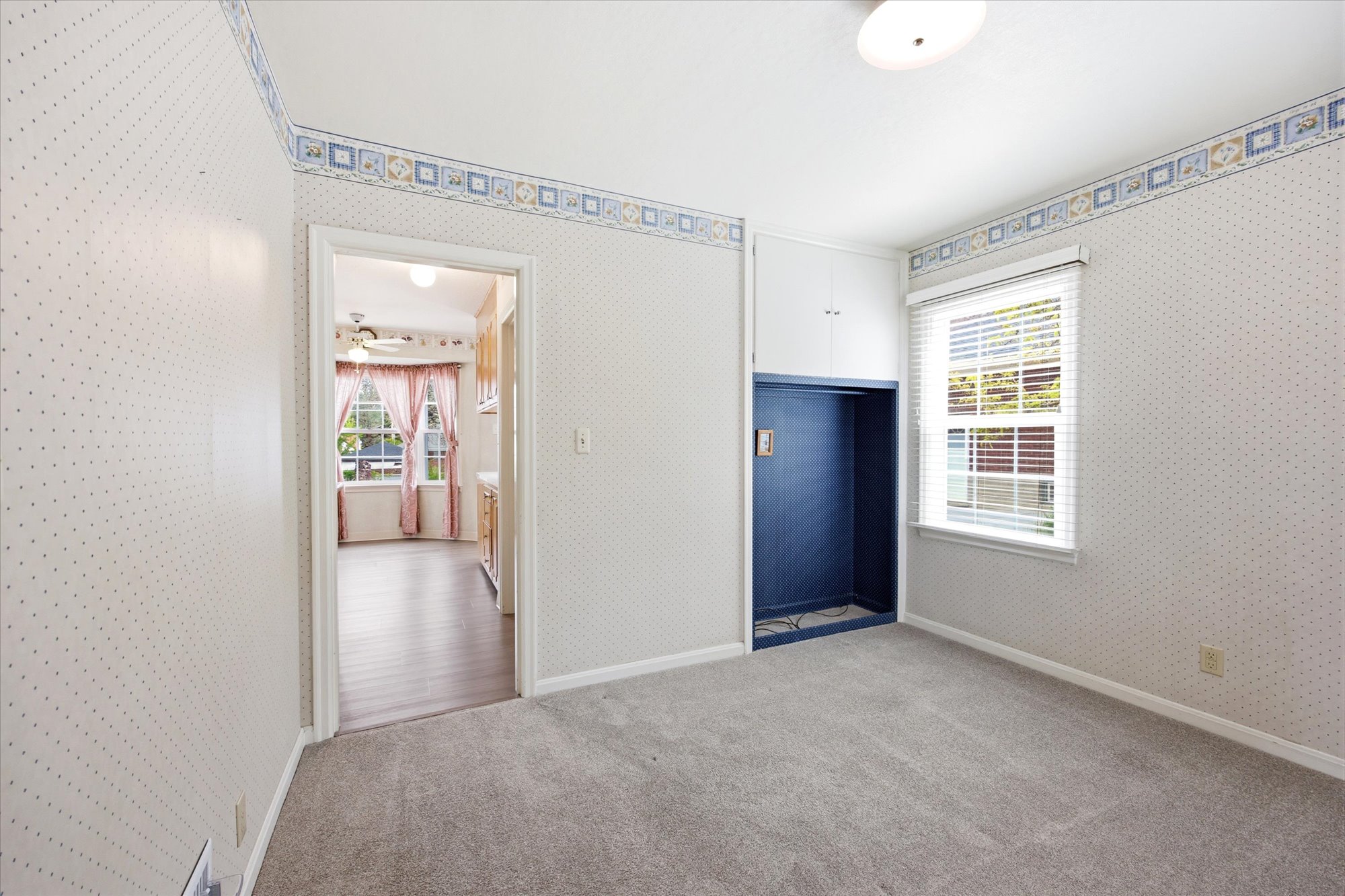 Carpeted interior space featuring a window with white blinds, built-in recessed shelving, and a decorative wallpaper border