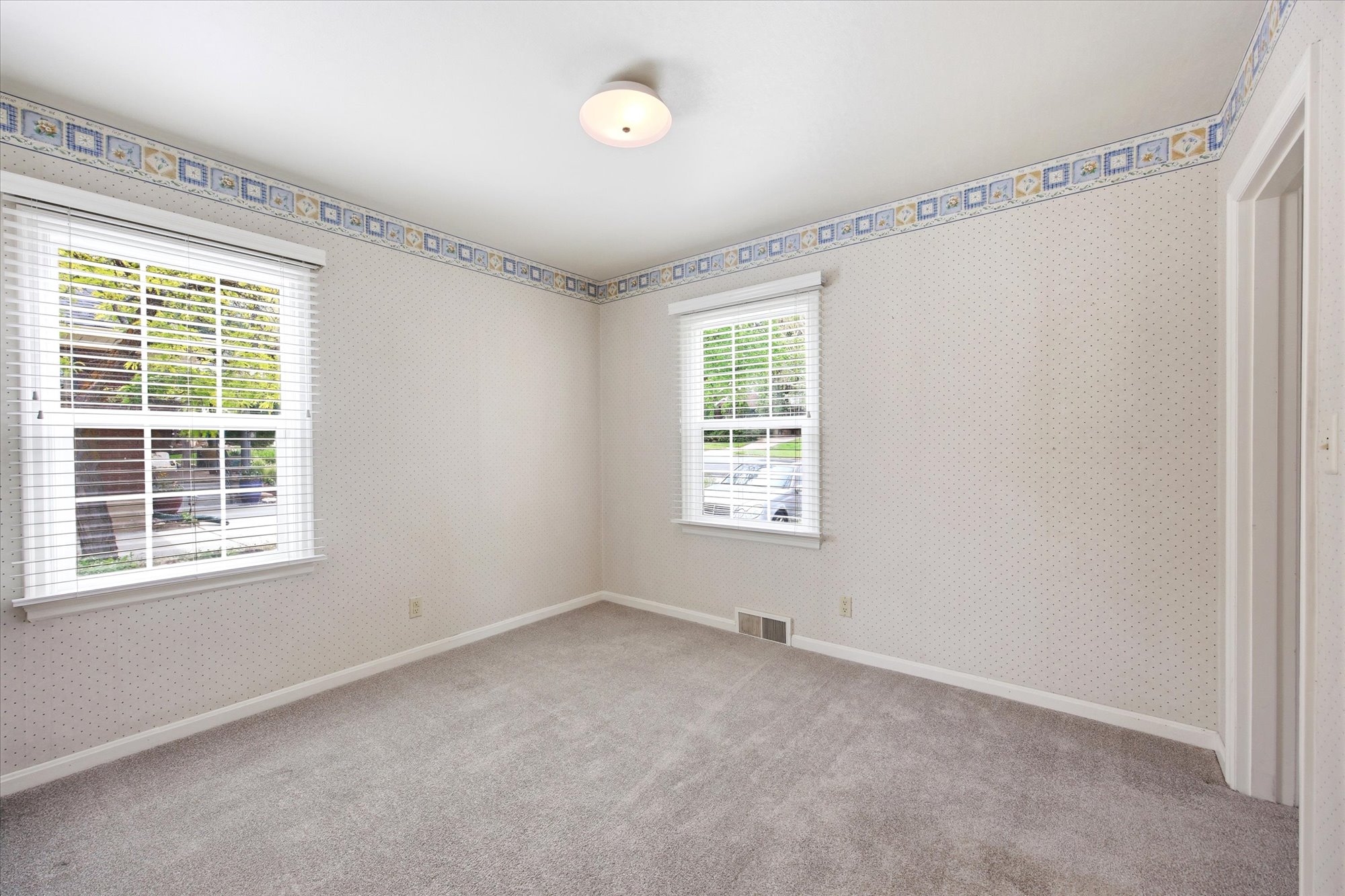 Carpeted room featuring two windows with horizontal blinds, patterned wallpaper, a decorative border, and a ceiling-mounted light fixture