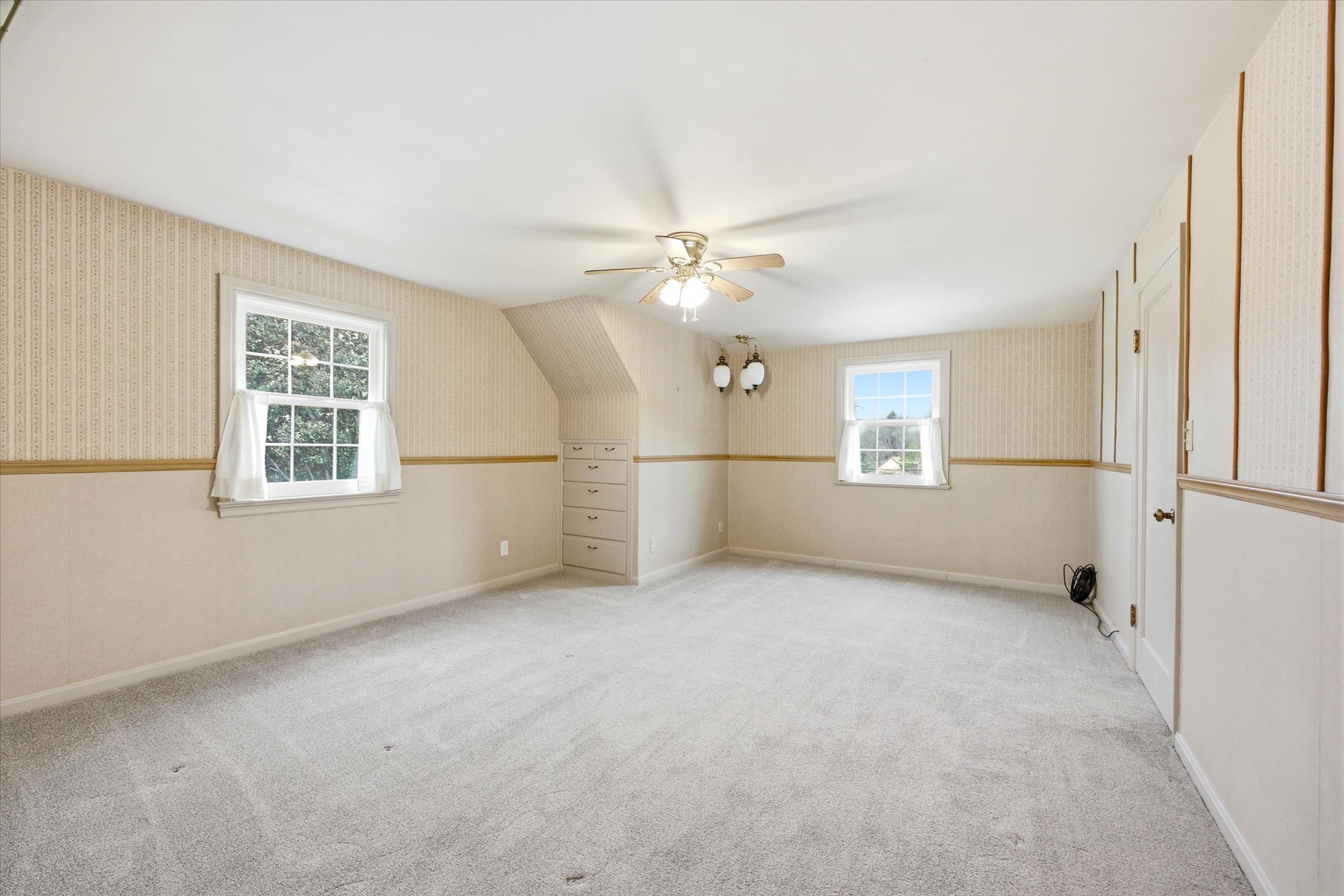 Spacious room featuring light-toned carpeting, two double-hung windows, a ceiling fan with integrated lighting, a built-in dresser with five drawers, and a wall-mounted light fixture