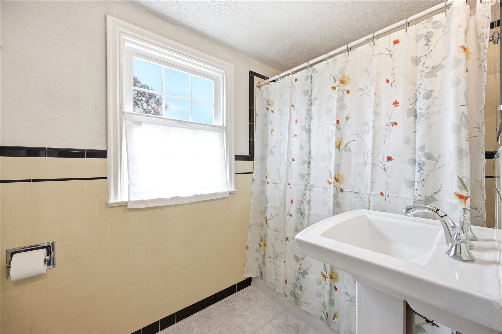 Pedestal sink with chrome faucet, light-toned wall tiling with black accent, and a single window with white trim