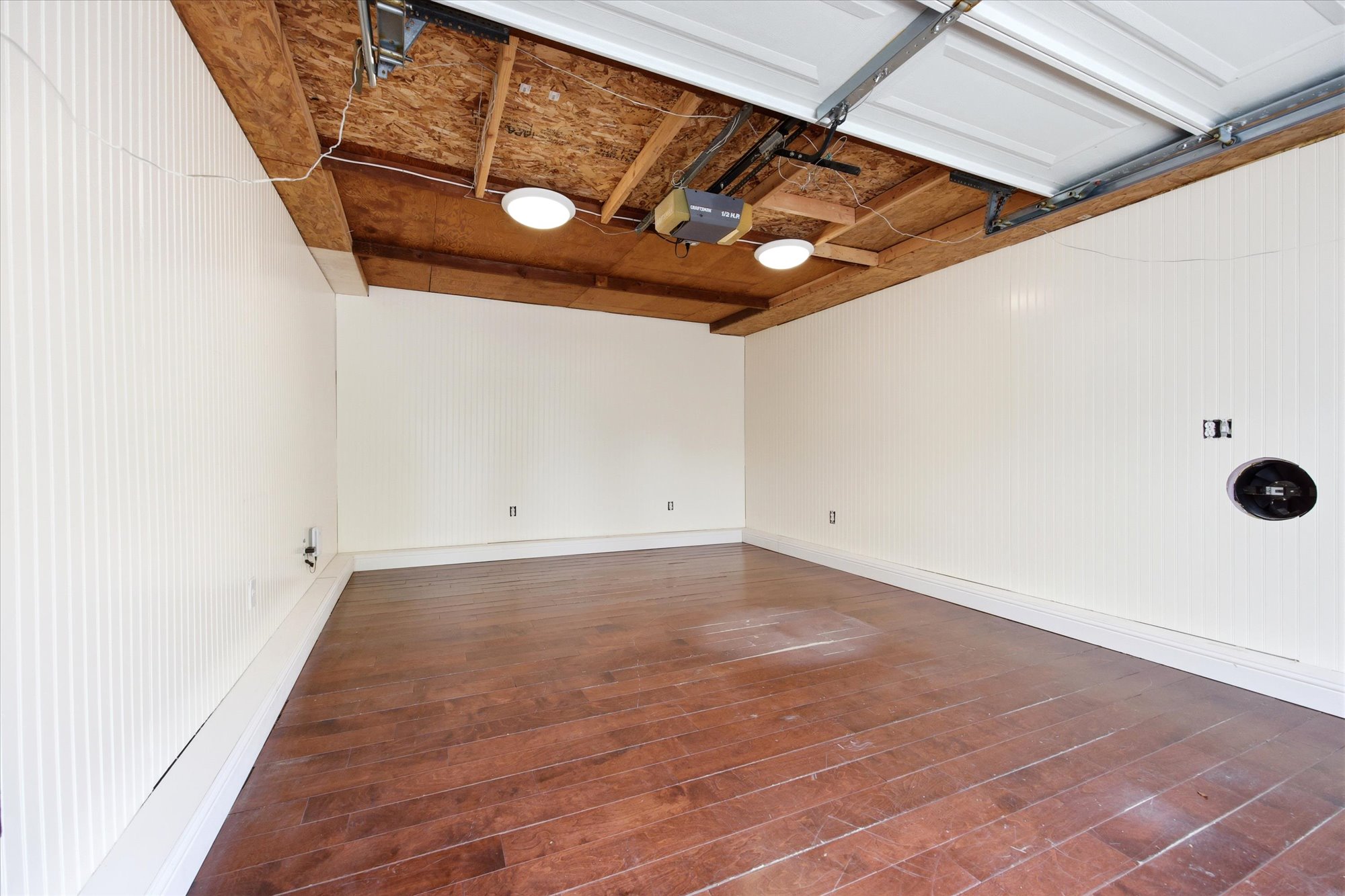 Garage interior featuring wood-finish flooring, beadboard wall paneling, and an exposed ceiling with integrated lighting