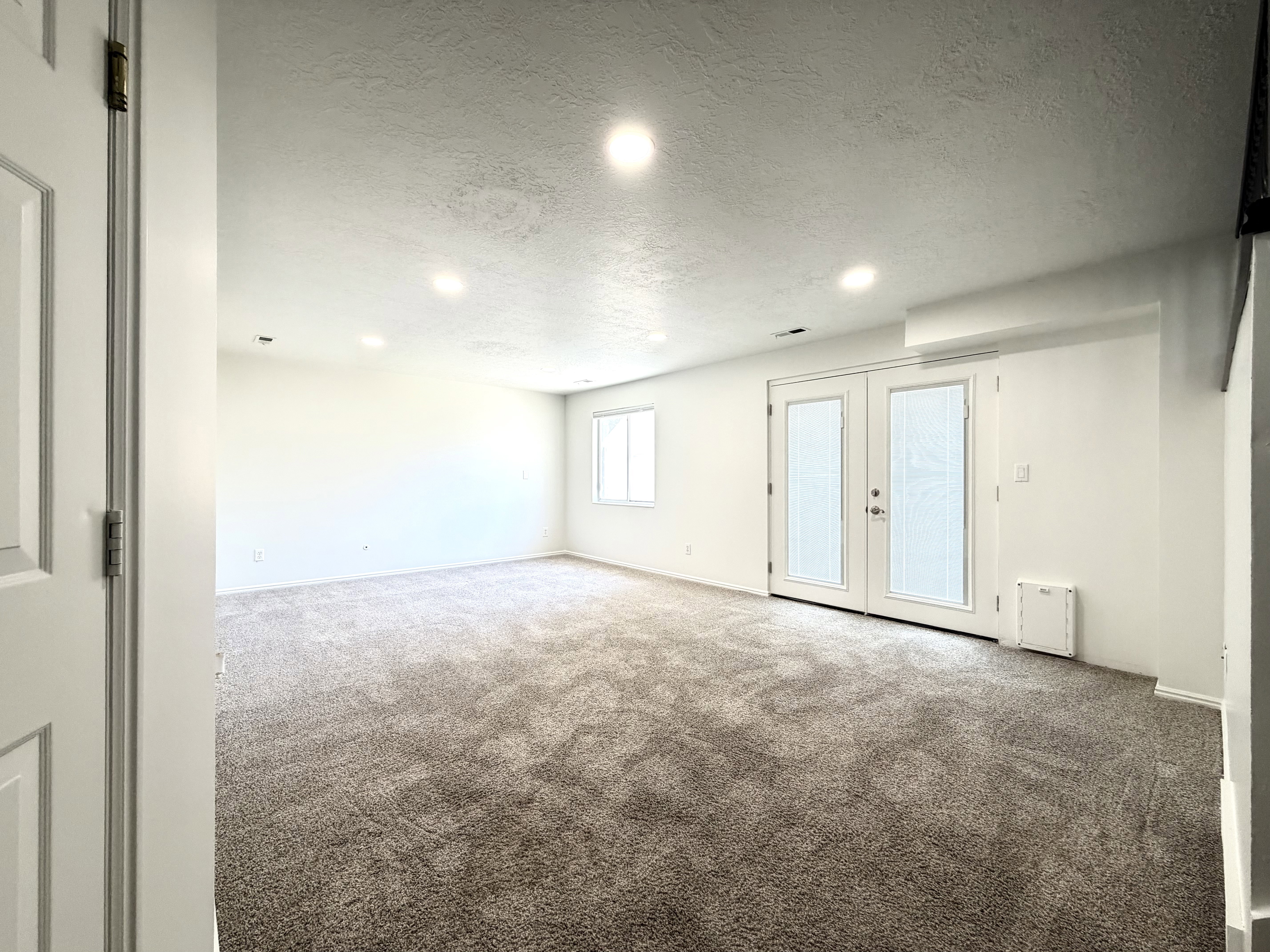 Spacious interior room featuring plush gray carpeting, white walls, recessed ceiling lighting, a dual window unit, and a set of French doors with integrated blinds