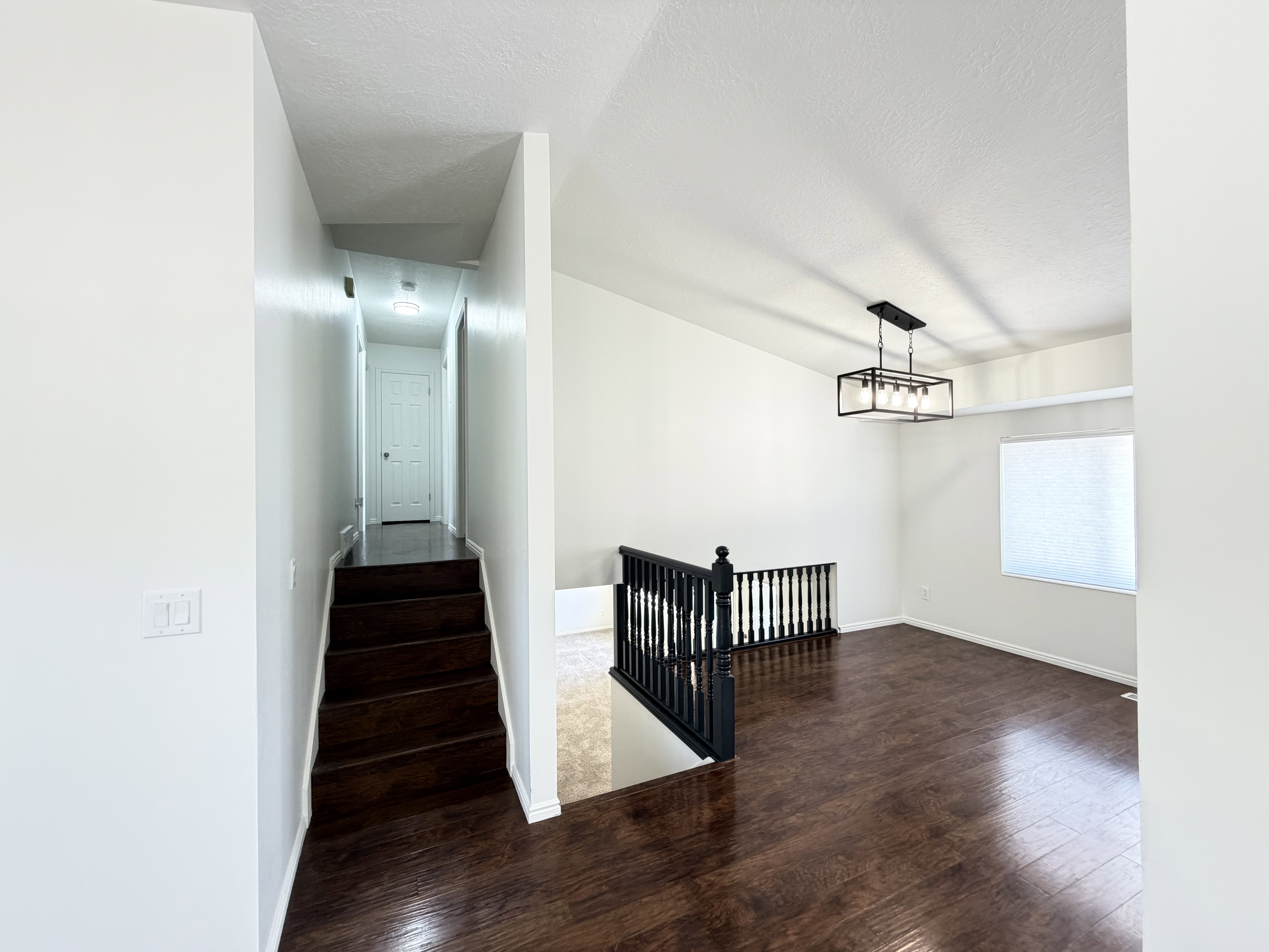 Spacious common area featuring rich wood-finish flooring, a modern linear chandelier, and a bright window with blinds