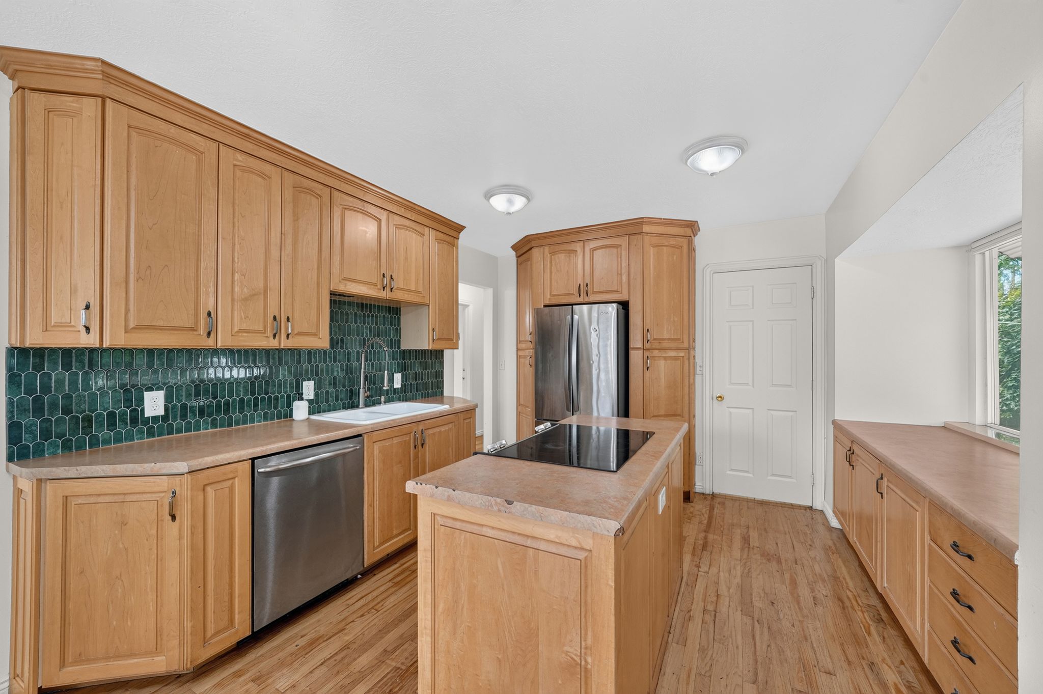 Kitchen featuring natural wood cabinetry, a striking green geometric tile backsplash, a center island with a built-in cooktop, stainless steel appliances, and wood-finish flooring