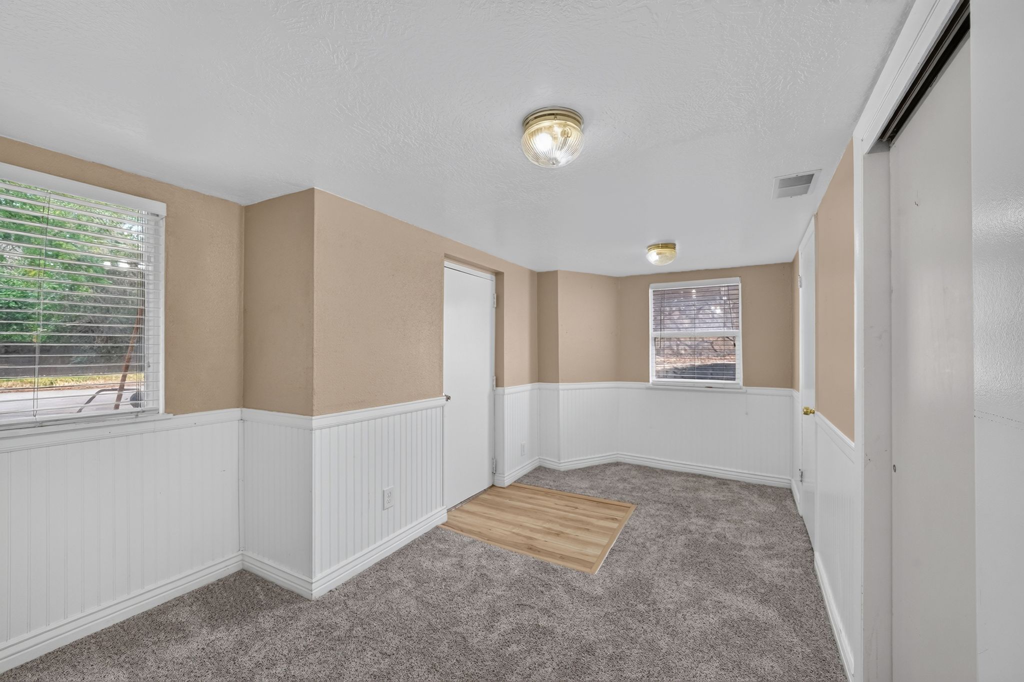 Inviting room featuring a window with horizontal blinds, neutral wall paint, white wainscoting, and gray textured carpeting with a wood-finish flooring insert