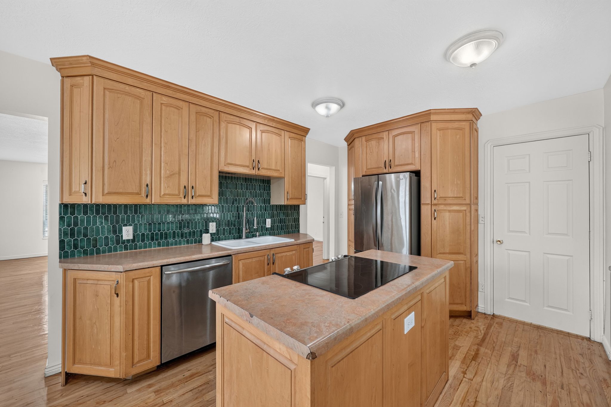 Kitchen featuring light wood cabinetry, a central island with an integrated cooktop, stainless steel appliances, and a green geometric tile backsplash