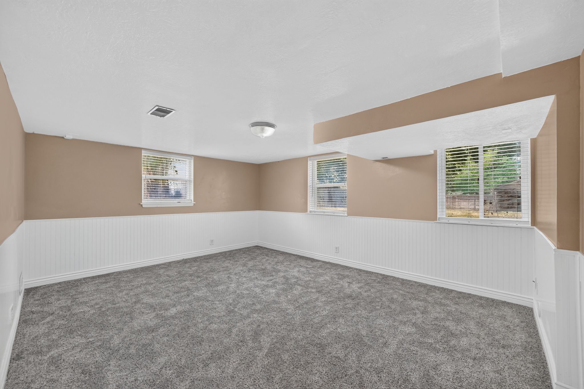 Spacious room featuring gray carpet flooring, white beadboard wainscoting, light brown walls, multiple windows with blinds, and recessed lighting