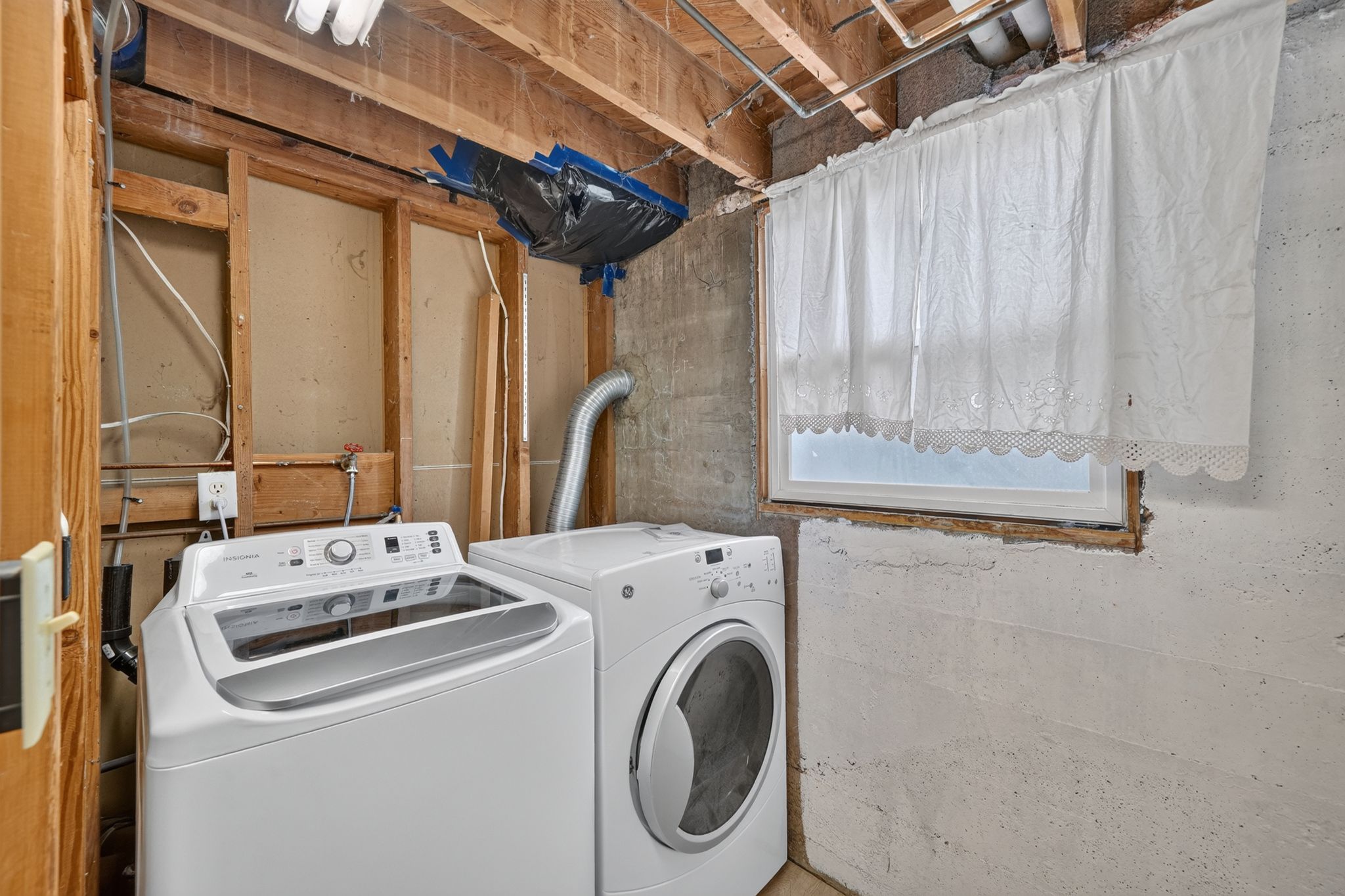 Utility area featuring a window with a white valance, exposed wood ceiling joists, and concrete walls