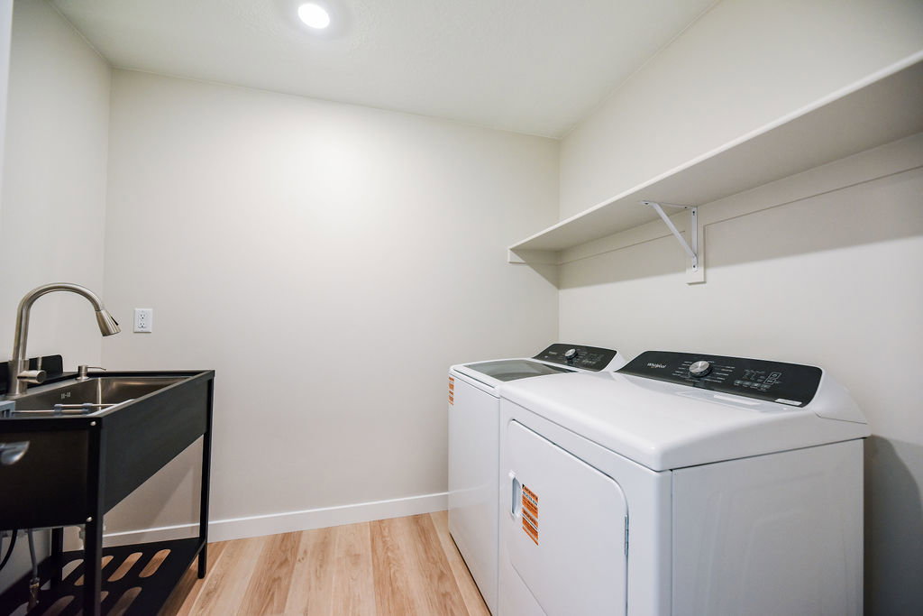 Dedicated laundry area featuring wood-finish flooring, a utility sink with a gooseneck faucet, a recessed ceiling light, and an overhead storage shelf