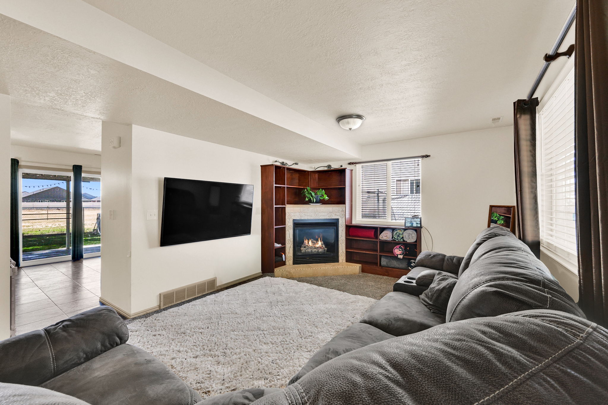Spacious living area featuring a corner fireplace with a stone surround, built-in shelving, and a large window with blinds