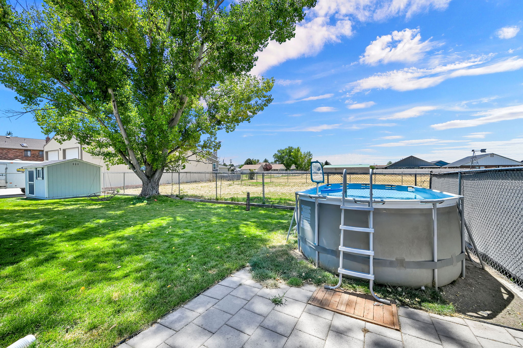 Expansive backyard featuring a mature shade tree, a manicured lawn, a chain-link fence, a paved patio area, and a storage shed