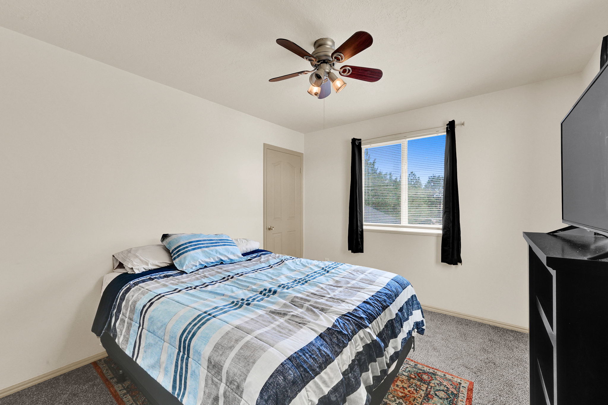 Carpeted bedroom featuring a ceiling fan with light fixture, a window with black curtains, and a light-colored interior door