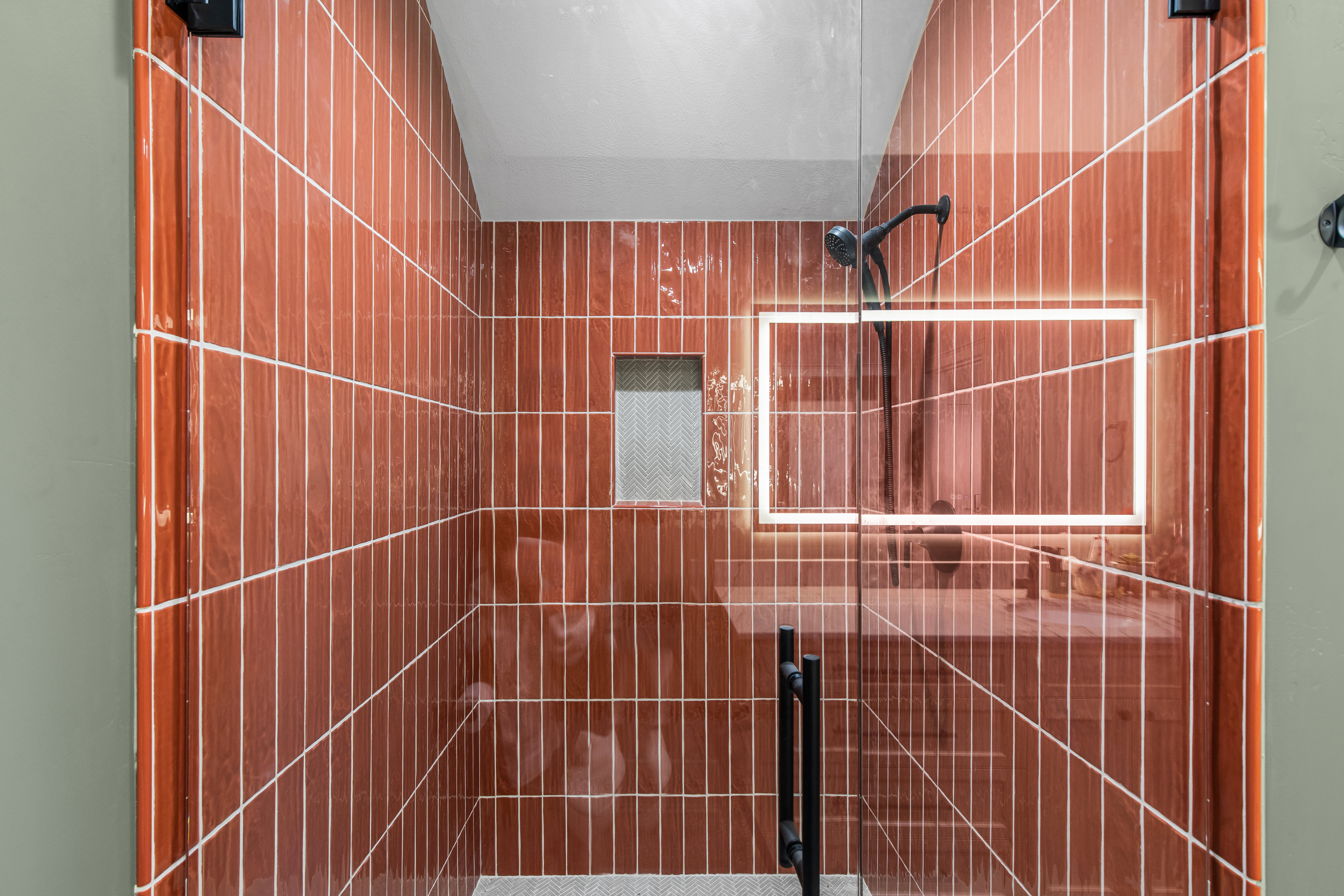 Glass-enclosed shower featuring vertically-stacked terracotta-hued wall tiles, black matte fixtures, and a recessed tiled niche