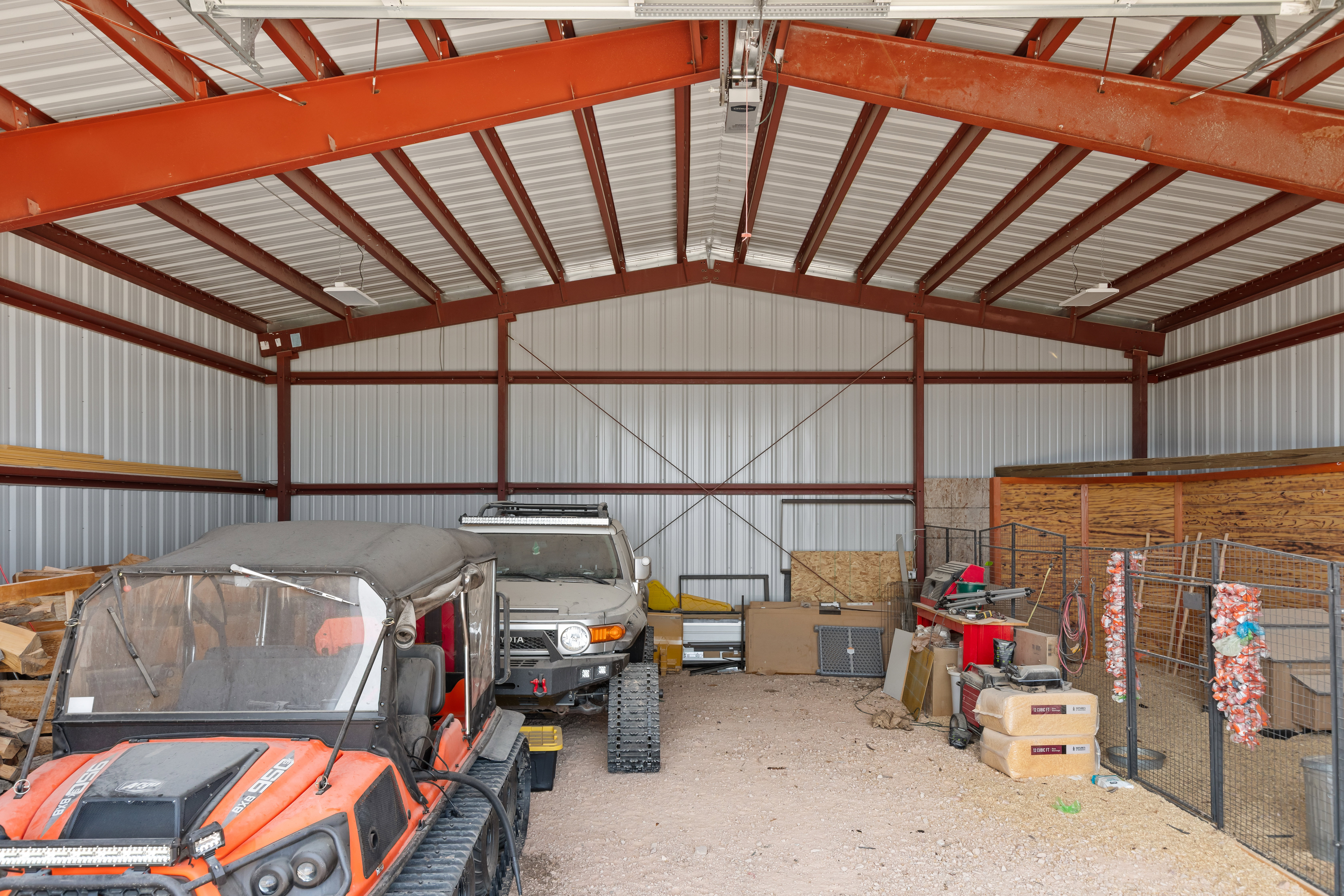 Spacious metal building featuring a vaulted ceiling with exposed red structural beams, corrugated metal walls, and a gravel floor
