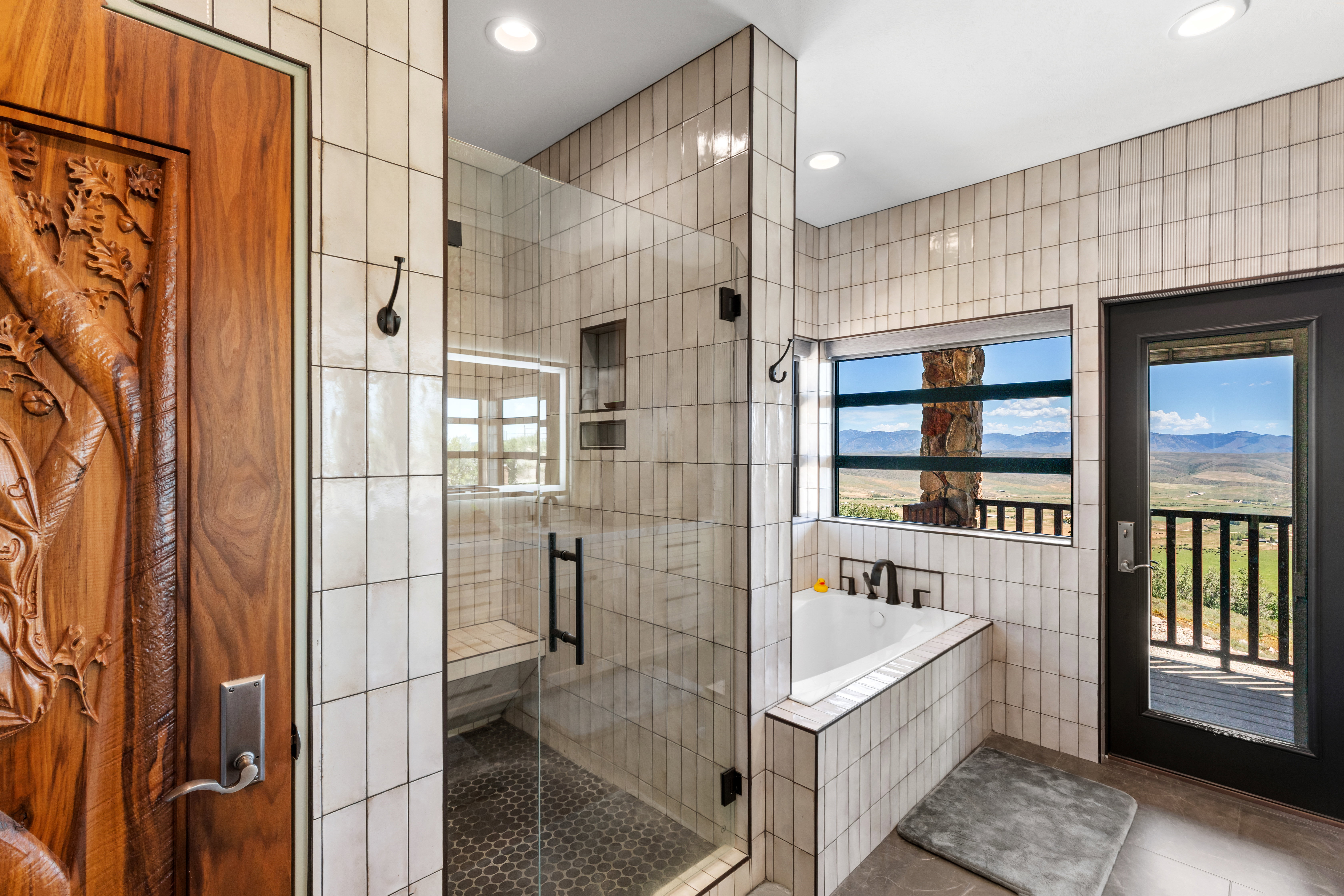 Bathroom featuring a clear glass shower enclosure, a built-in tub, white vertical tile accents, a private exterior balcony, and a carved wood door