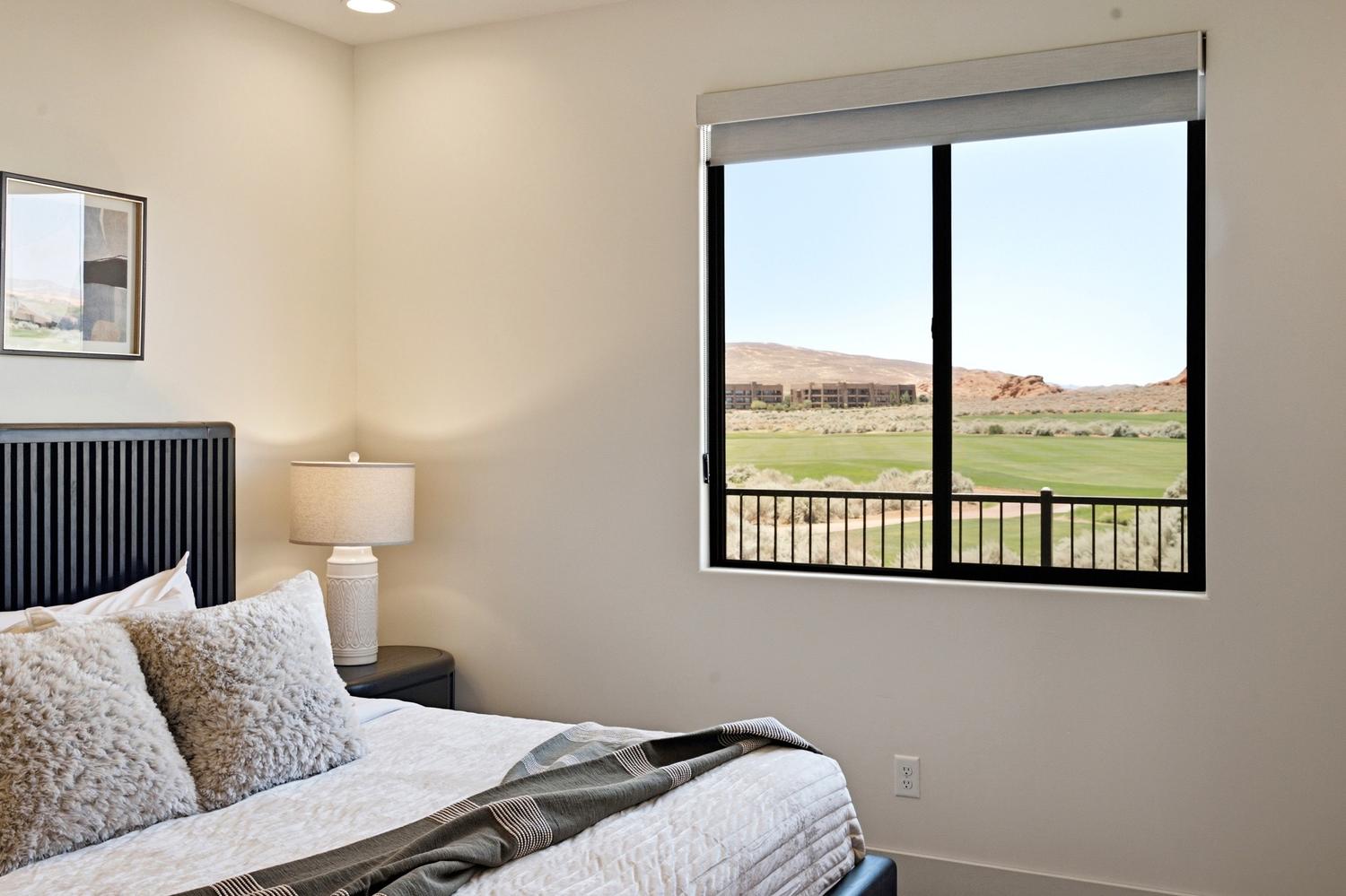 Bedroom featuring a large window with black framing, a roller shade, and a landscape with green terrain and distant structures