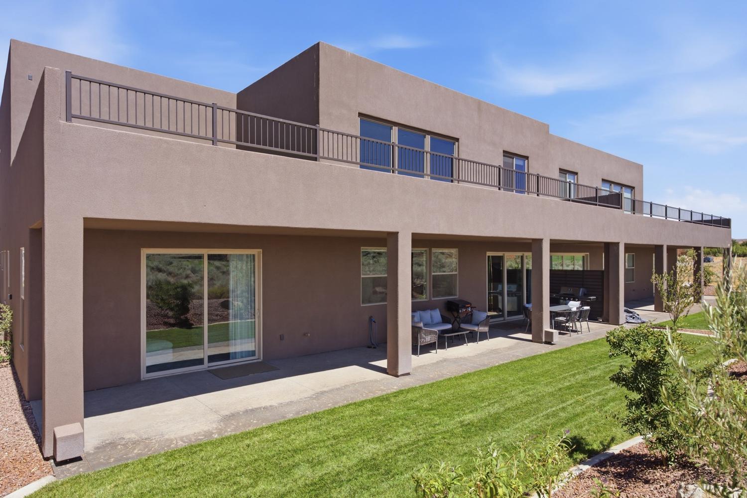Contemporary stucco exterior with a full-length covered patio, expansive upper-level balcony featuring metal railings, multiple sliding glass doors, and a manicured lawn