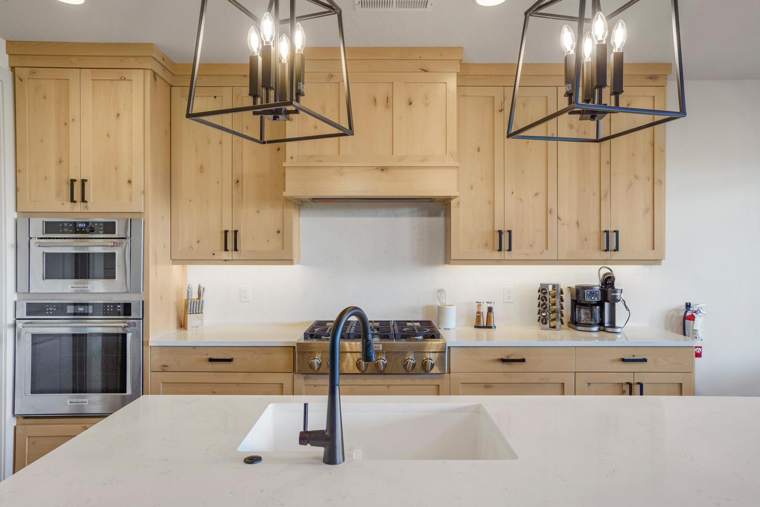 Kitchen featuring light wood-finish cabinetry with black hardware, stainless steel built-in double ovens, a gas range with a matching range hood, and an undermount sink with a black faucet
